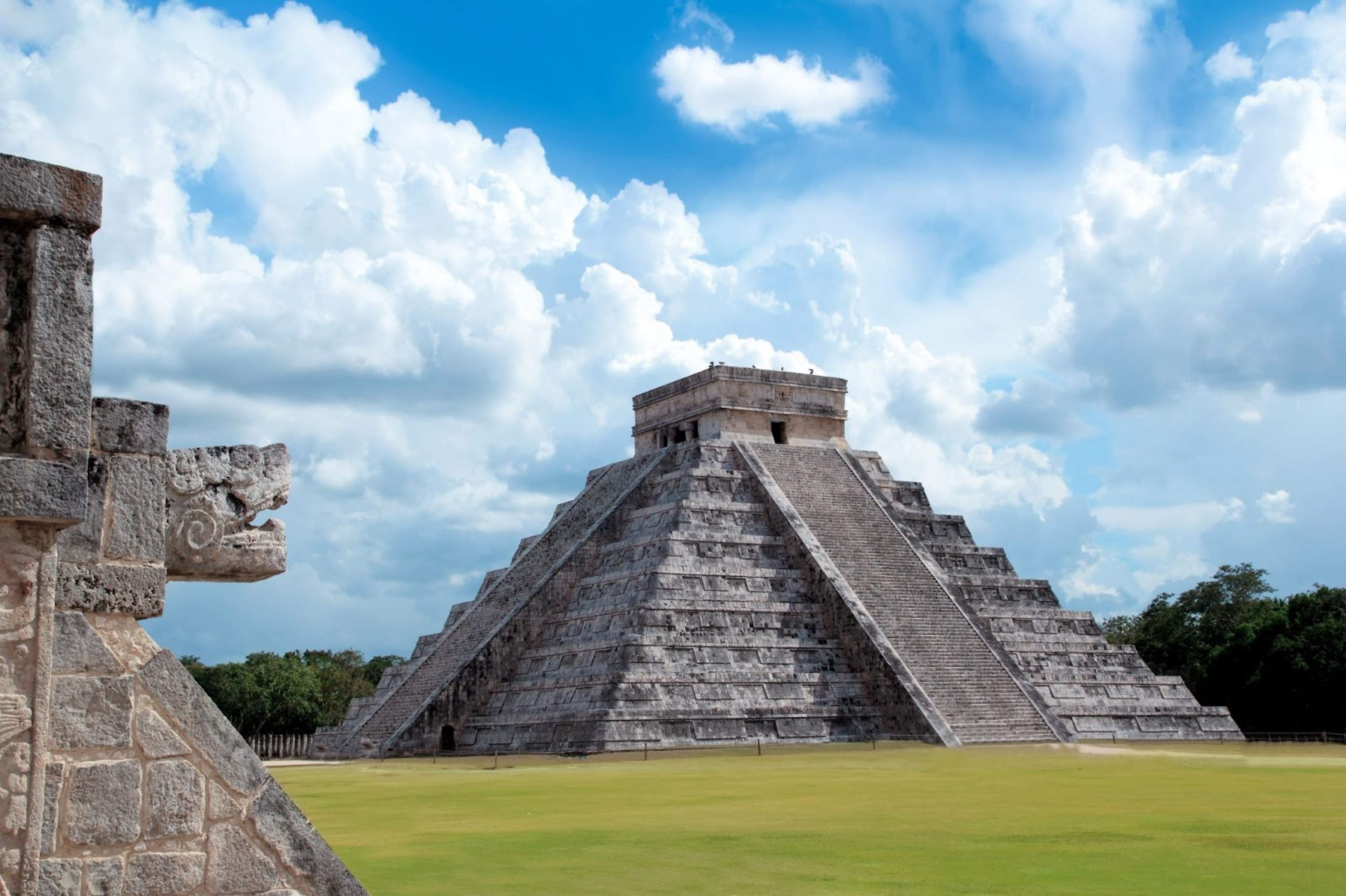 El Castillo, the pyramid at Chichen Itza, under a bright blue sky with clouds, featuring a stone serpent head in the foreground.