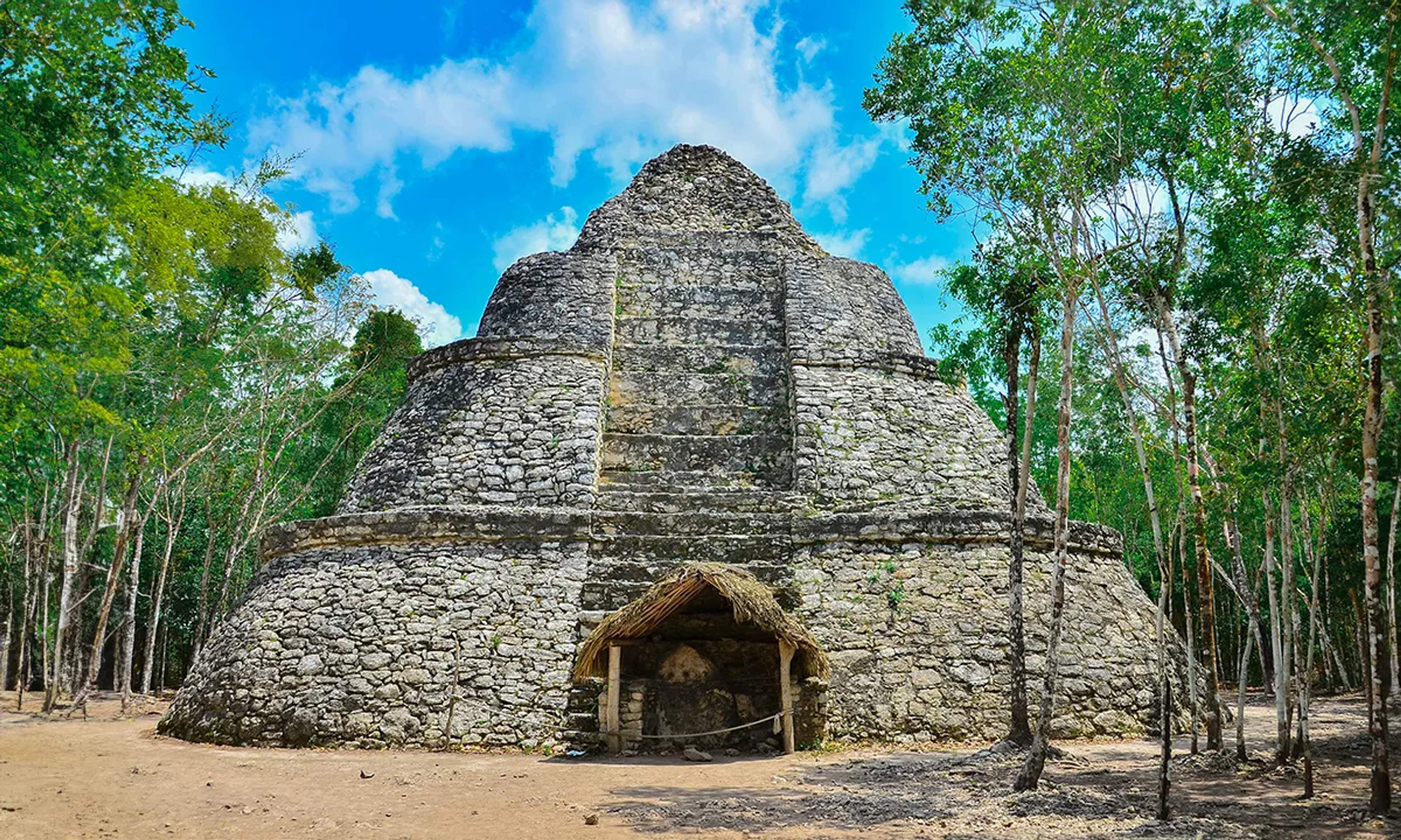 Pirámide circular de piedra con una choza de techo de palma en su base, rodeada de árboles y un cielo azul brillante.