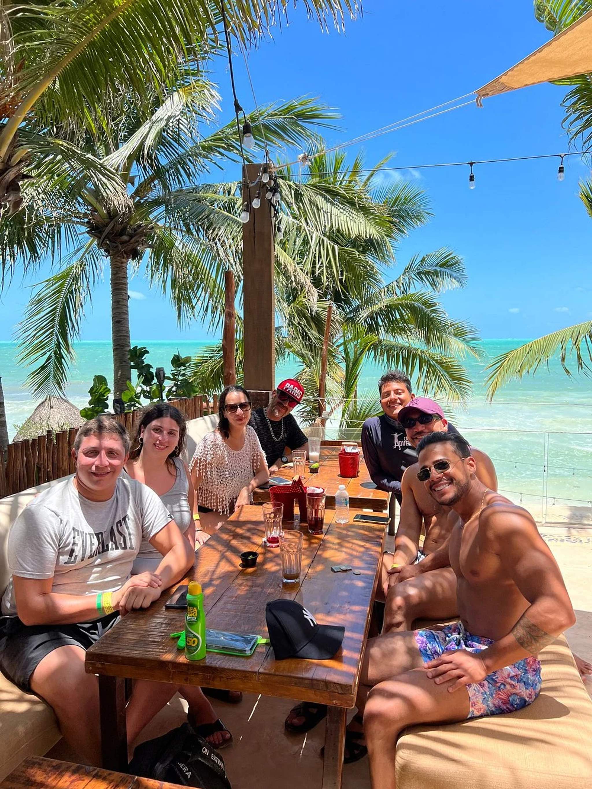  Un grupo de amigos disfrutando de un día soleado en un restaurante frente a la playa en Cancún, con palmeras y el océano de fondo.