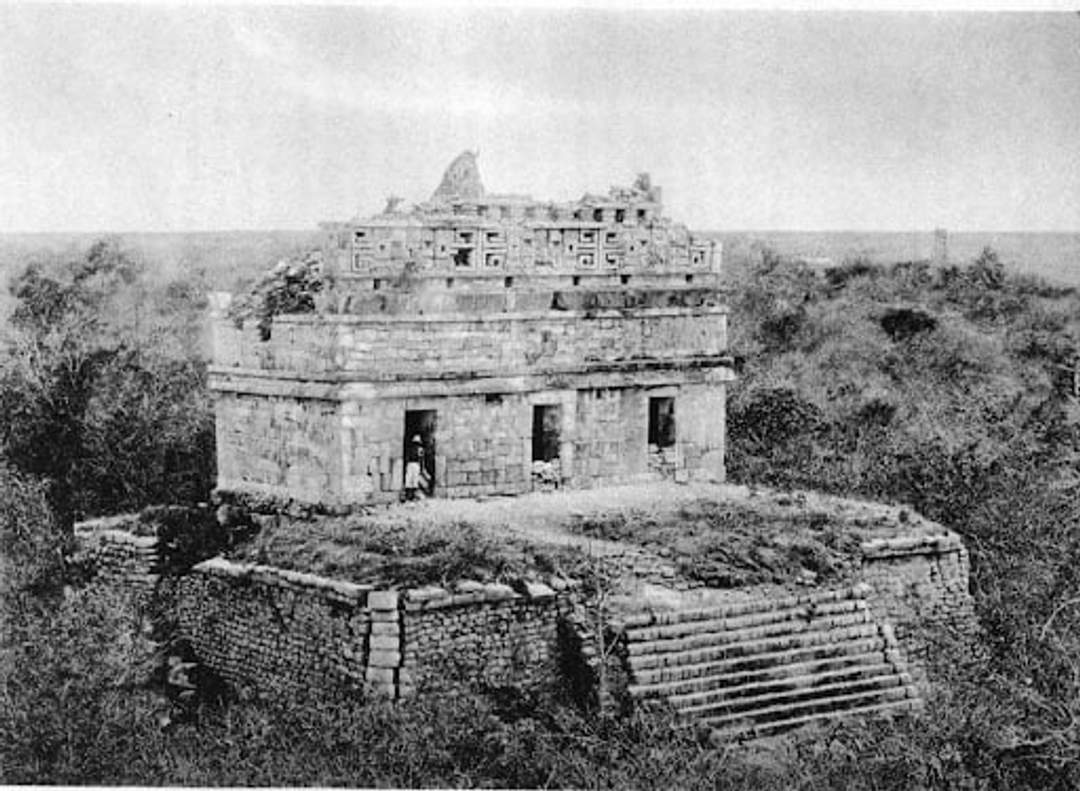 Foto en blanco y negro de un edificio antiguo de piedra con intrincados grabados en la sección superior, rodeado de densa vegetación. Una escalera conduce a la estructura elevada, sugiriendo un sitio histórico o arqueológico.