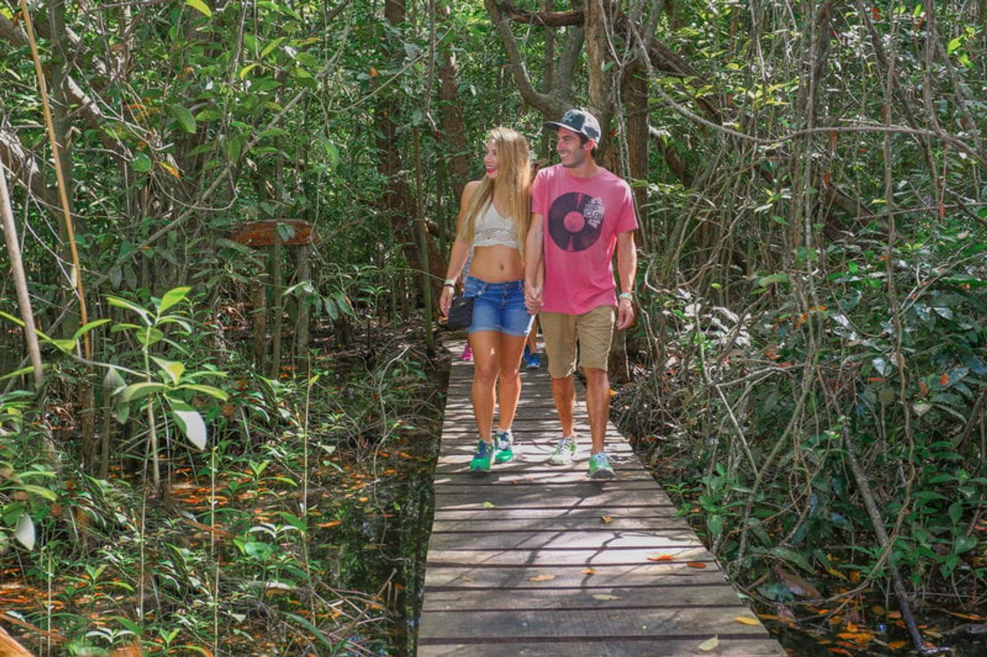 A couple holding hands and walking on a wooden path through lush greenery in a tropical forest.