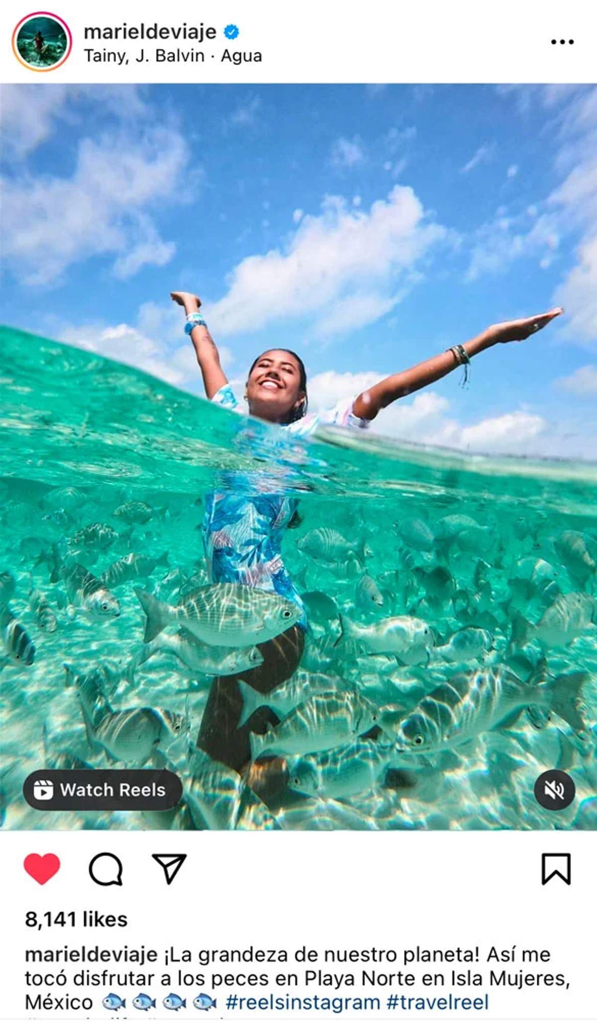 Una mujer con un traje de baño azul está parcialmente sumergida en agua turquesa clara, rodeada de peces.