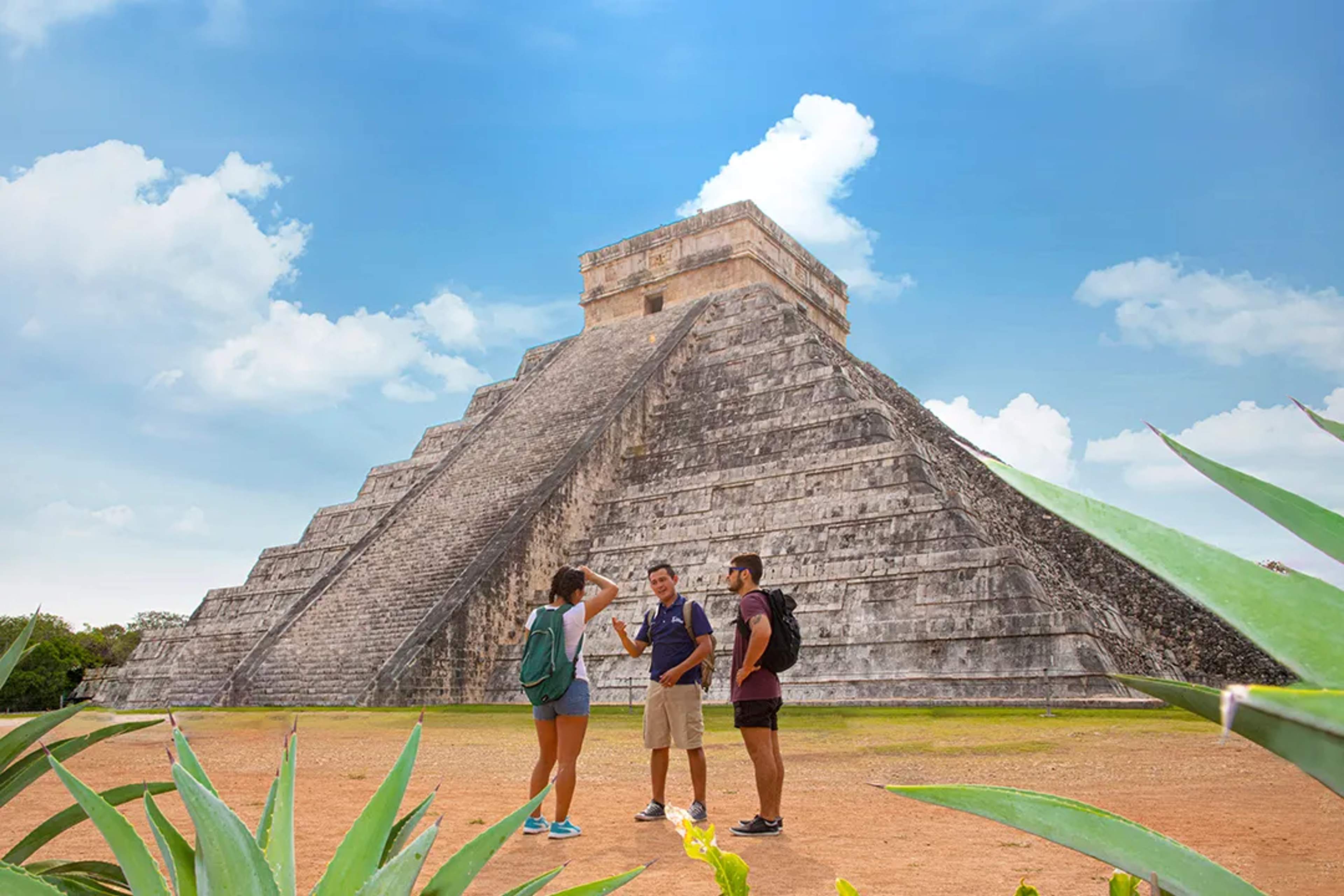 Tres turistas en la pirámide de Chichén Itzá, Cancún, hablando sobre su historia bajo un cielo despejado.