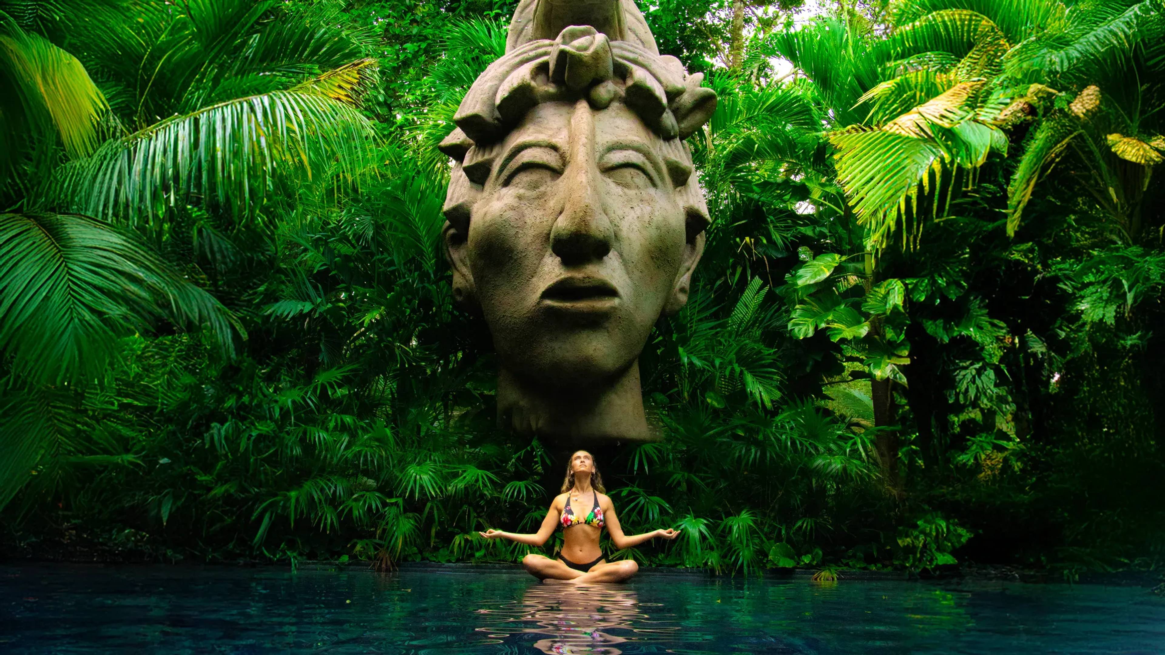 Woman meditating in a pool surrounded by lush jungle, under a large Mayan statue in Tulum.