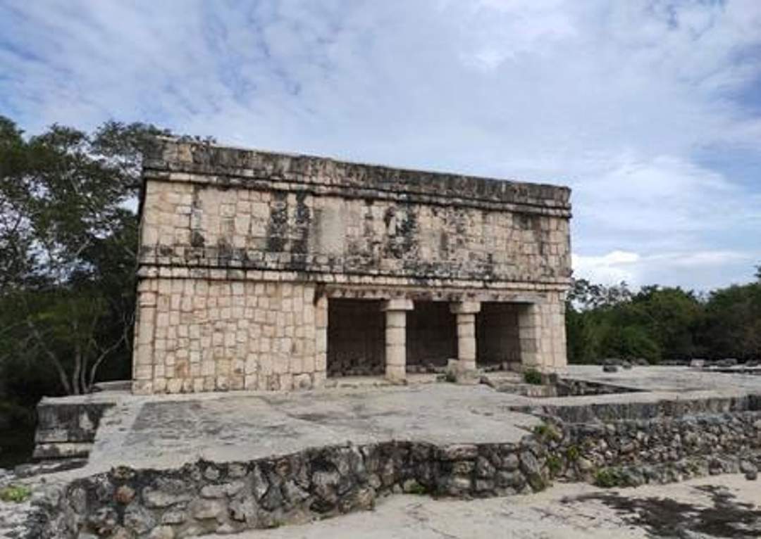 A photograph of the Temple of the Dancing Jaguars at Chichen Itza, featuring a rectangular stone building with two columns supporting a central entrance. The structure is elevated on a stone platform and surrounded by trees. Photo credit: Giovanni Agostino Frassetto.