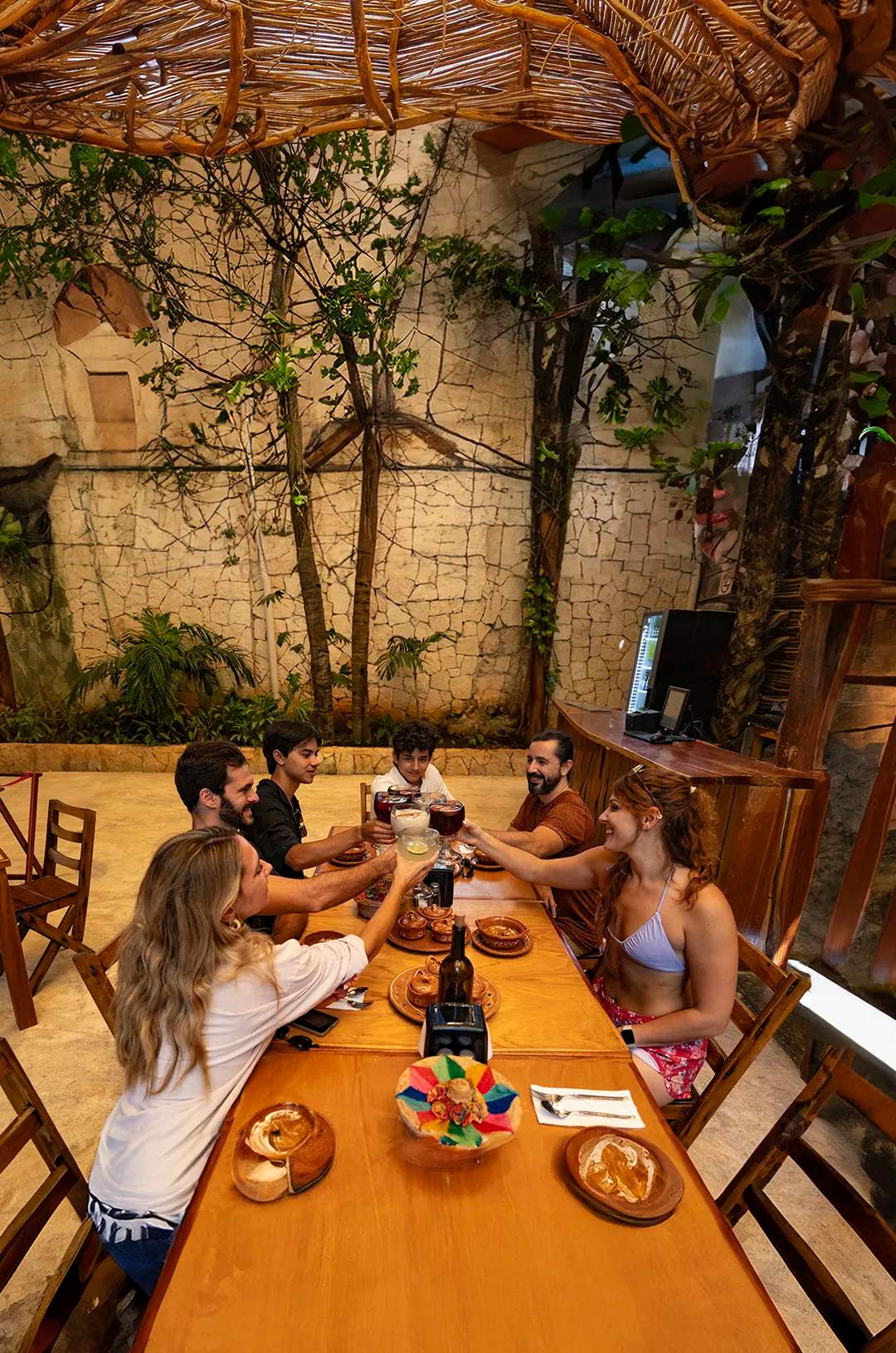 Un grupo de personas sentadas alrededor de una mesa de madera en un restaurante rústico, brindando con bebidas y sonriendo.