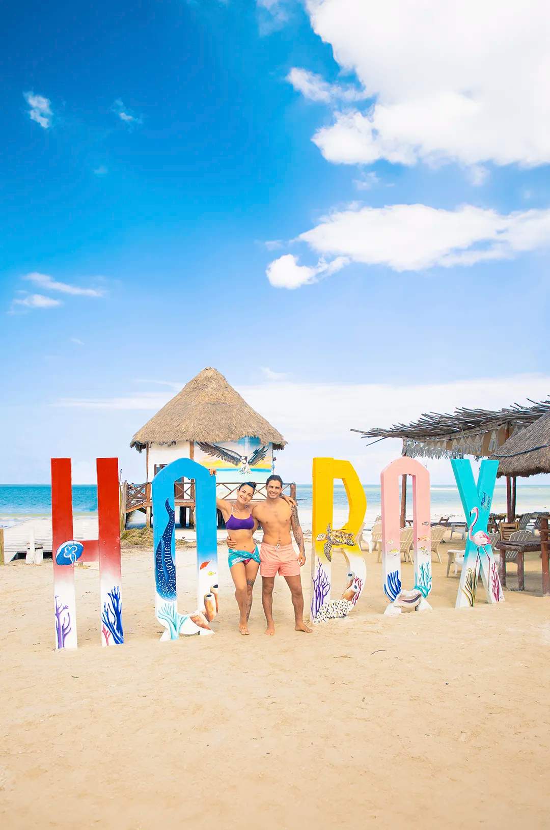 Couple in swimwear posing by colorful "Holbox" sign on the beach with ocean and thatched huts in the background