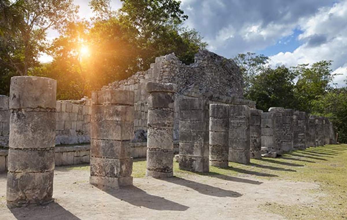 Ancient stone columns and ruins at Chichen Itza, with sunlight peeking through trees in the background.