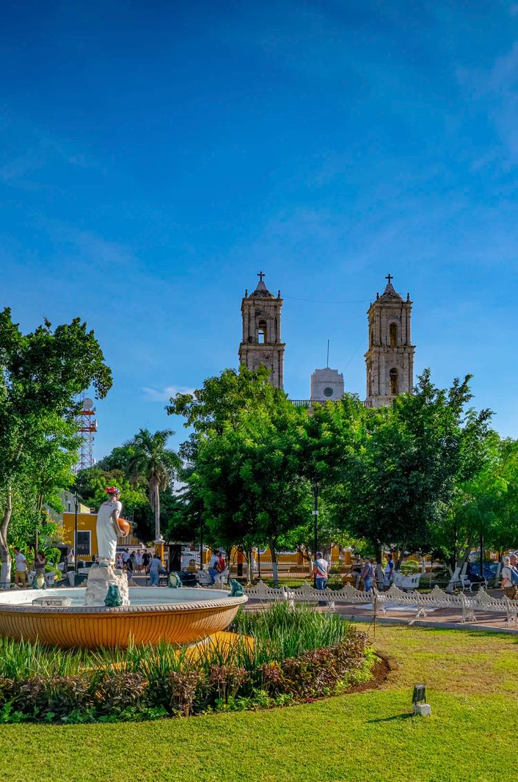 Main square of Valladolid with the fountain and San Gervasio Cathedral in the background, surrounded by trees.