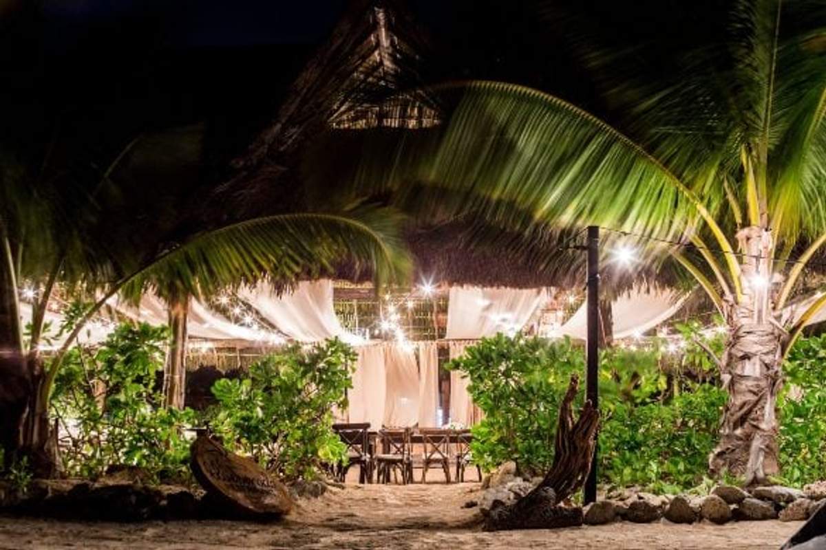 A cozy beachside restaurant at night, adorned with string lights and nestled among palm trees and lush greenery. The ambiance is warm and inviting, with softly lit tables set under a thatched roof.