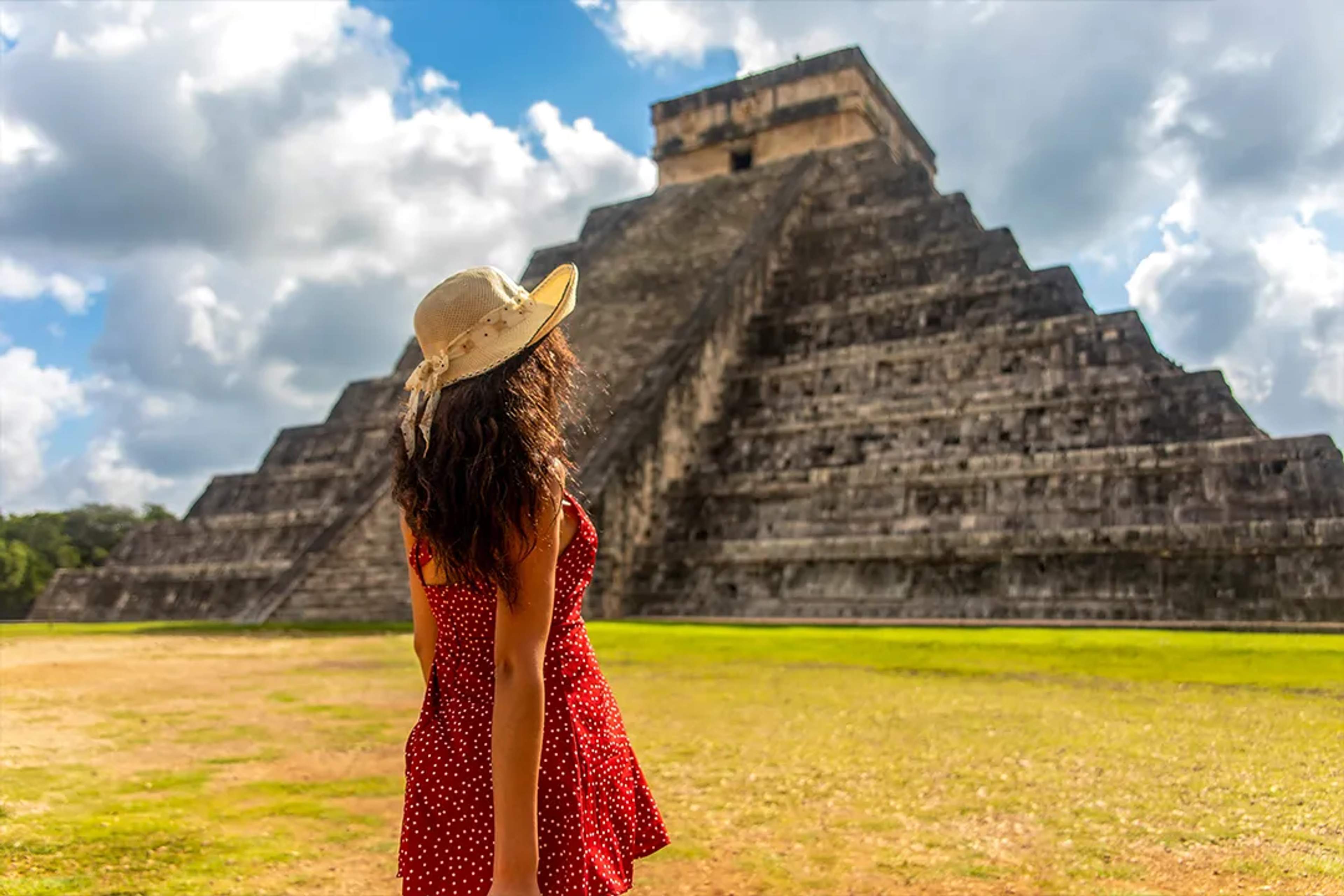 Mujer con sombrero admira la antigua pirámide maya de Chichen Itzá bajo un cielo parcialmente nublado.