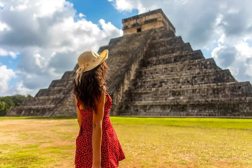 Woman in a hat admires the ancient Mayan pyramid at Chichen Itza under a partly cloudy sky.