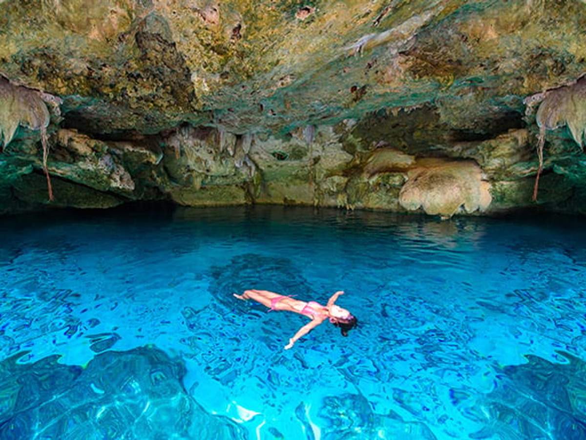 Person swimming in a cenote with clear blue water, surrounded by natural cave formations.