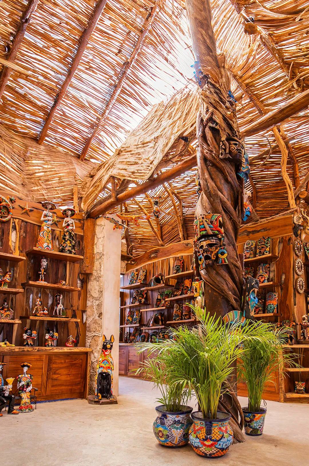 Tienda rústica con techo de paja, cerámica colorida, figuras y máscaras en estantes de madera, y plantas en macetas.
