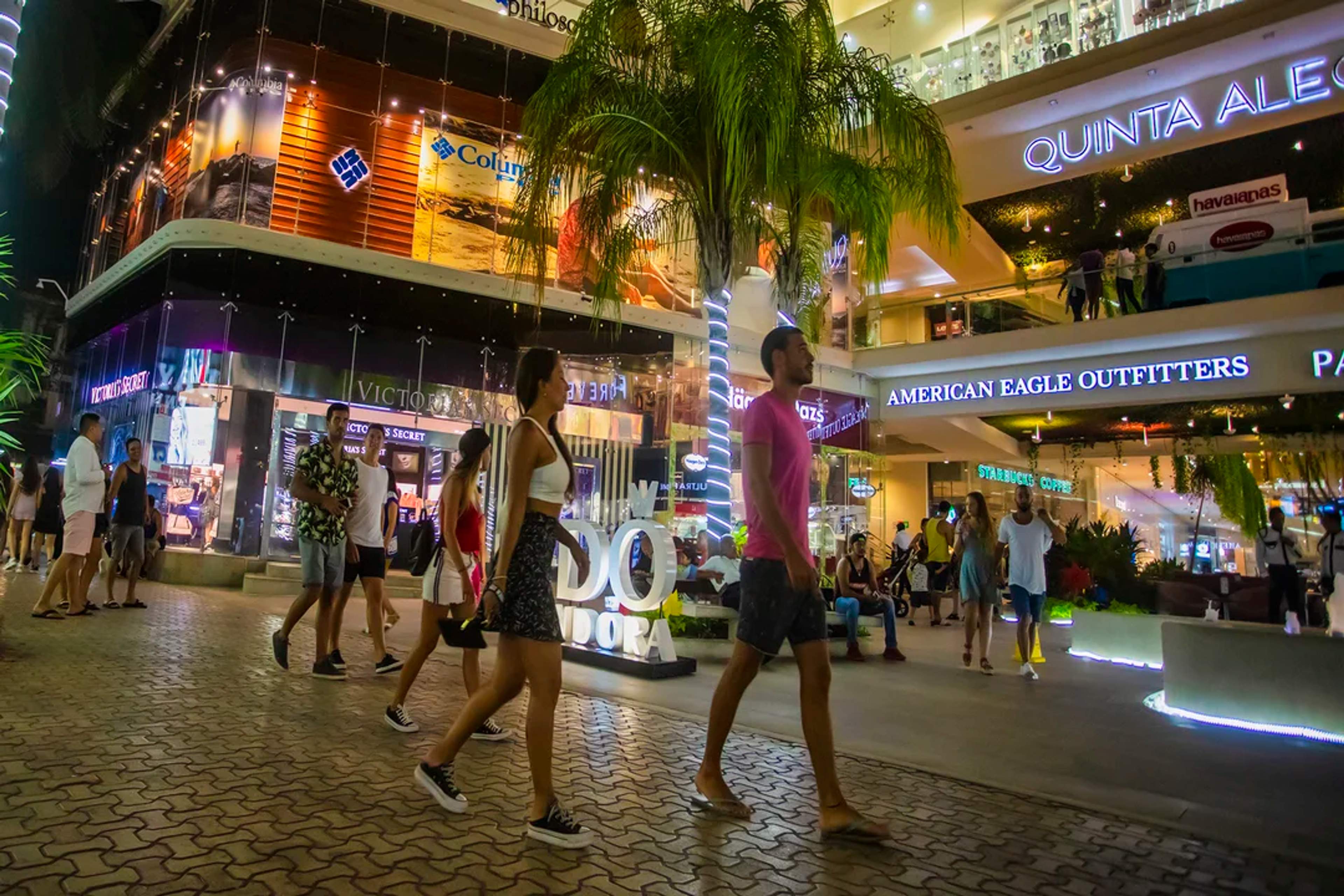 Turistas pasean por una animada zona comercial nocturna, rodeados de tiendas y luces brillantes.