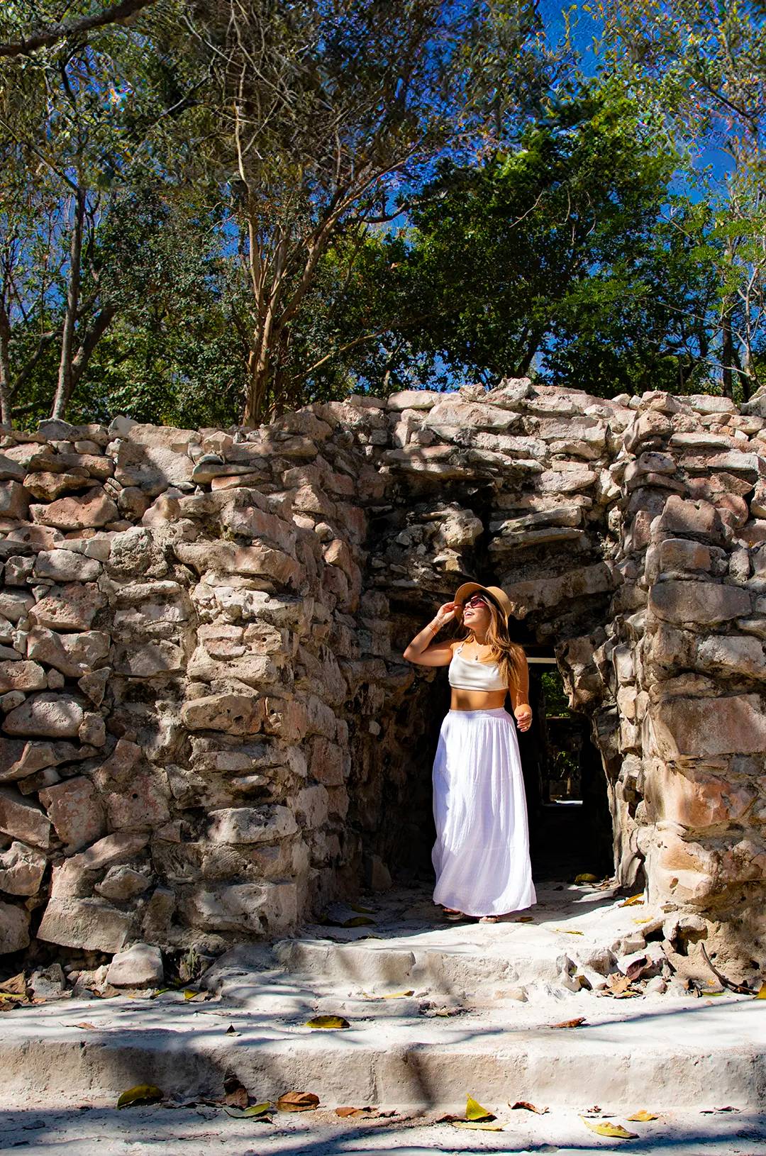 A woman stands in a stone archway at an ancient ruin site, wearing a hat and a white skirt, enjoying the sunlight.