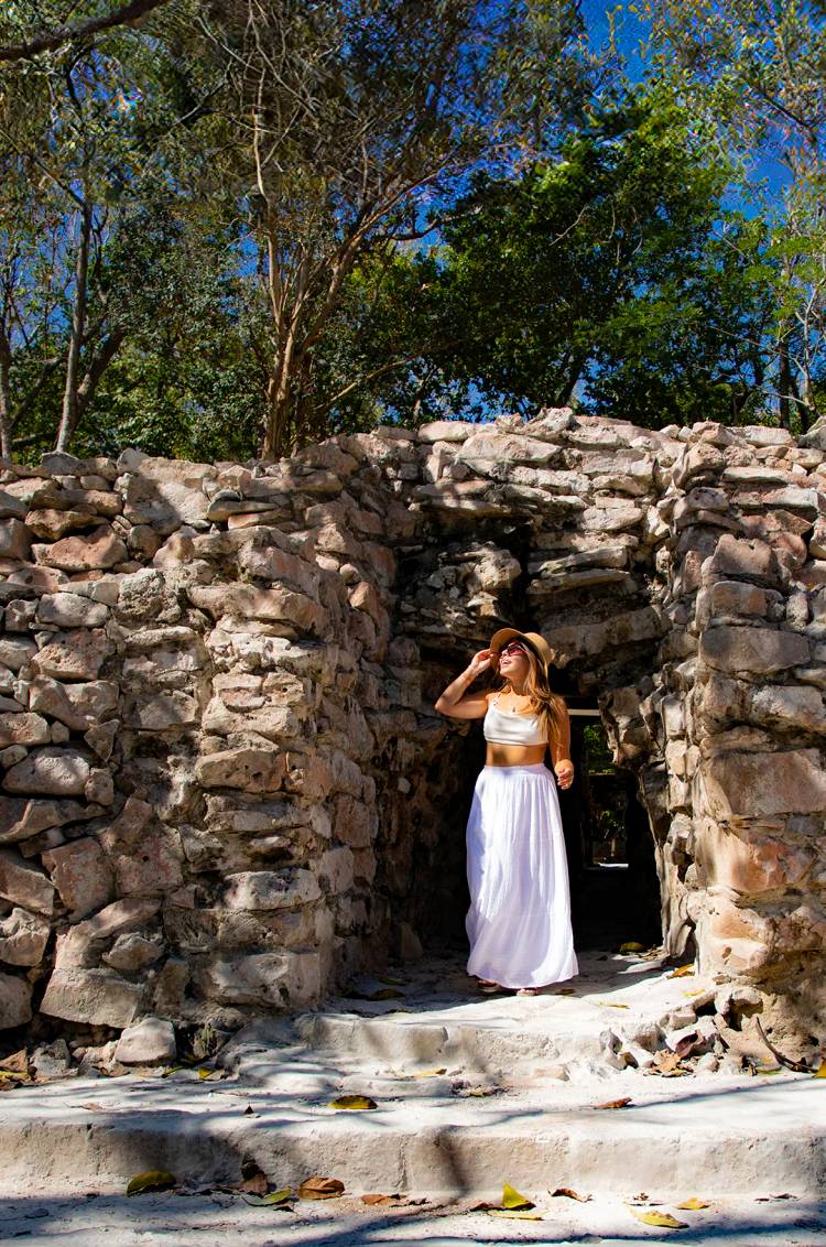 Mujer en un arco de piedra en un sitio de ruinas antiguas, con sombrero y falda blanca, disfrutando de la luz del sol.