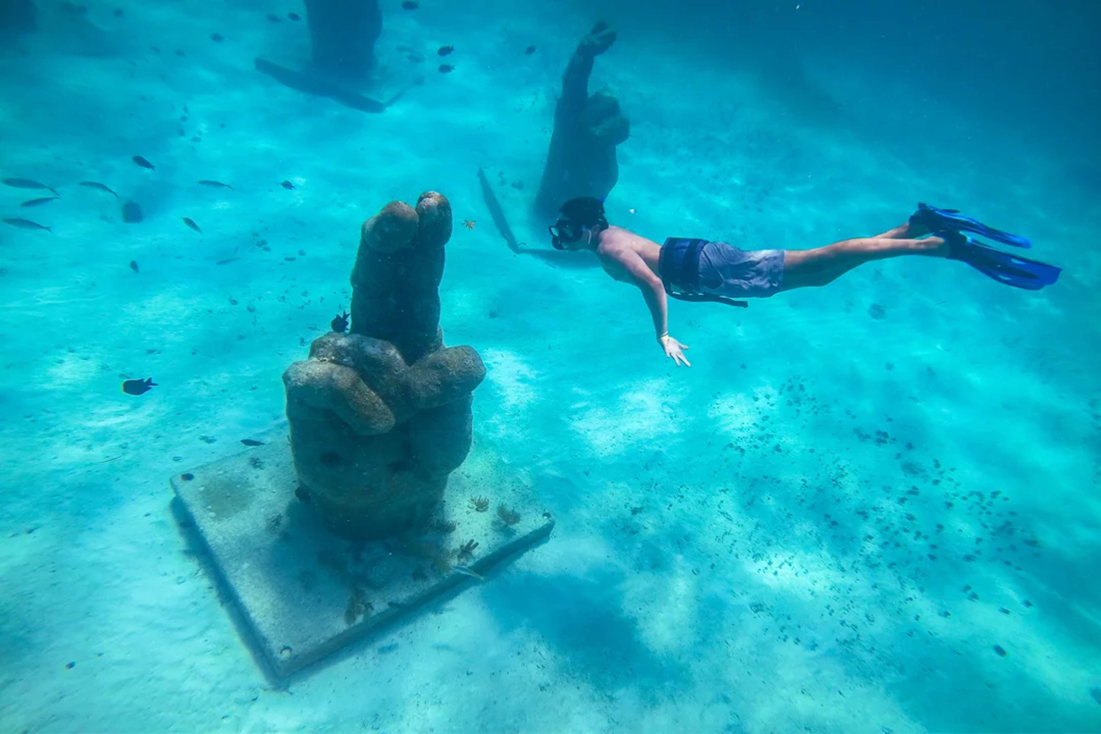 Snorkeler explores underwater sculptures in clear blue waters surrounded by tropical fish.