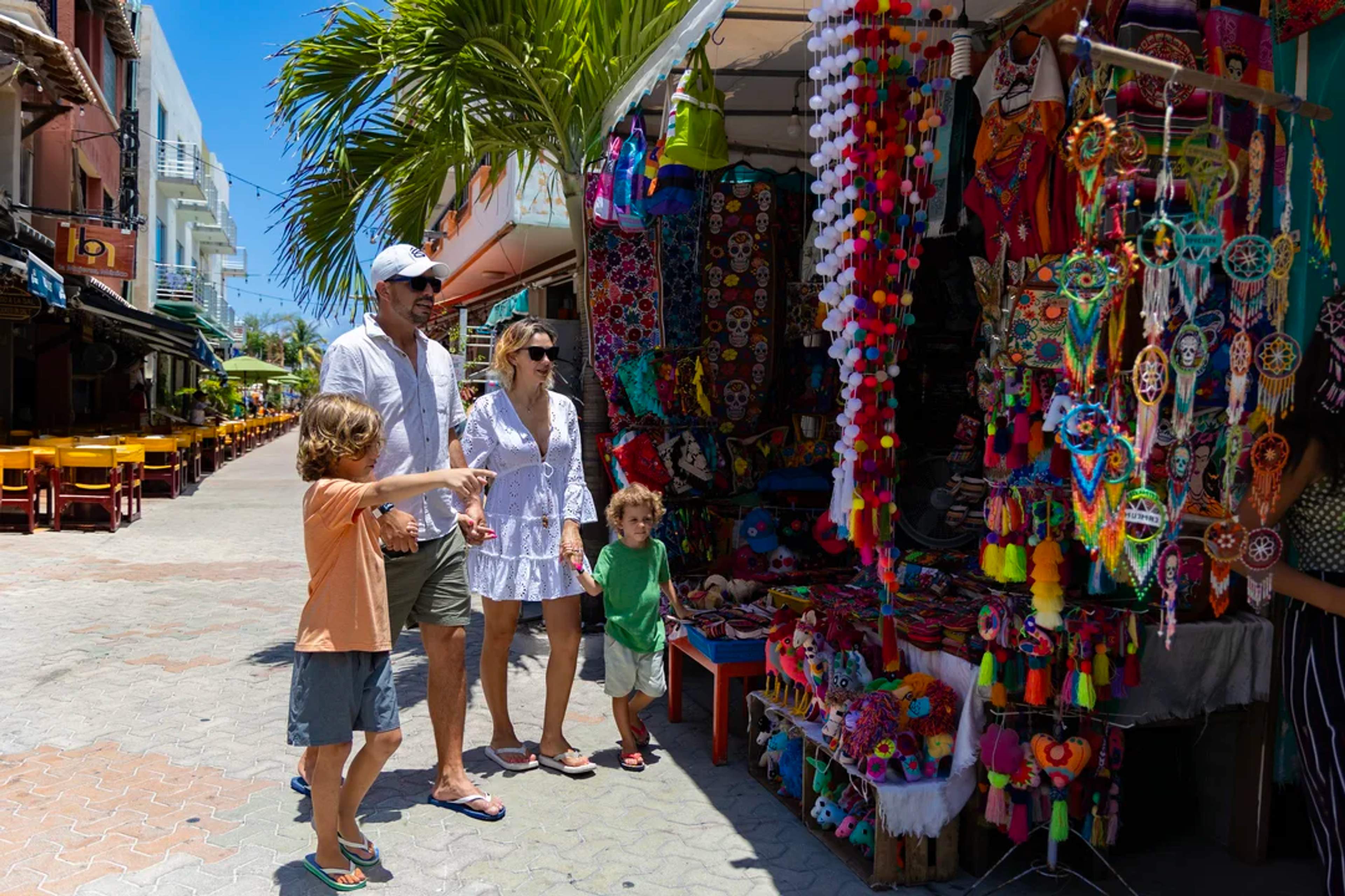 Una familia recorre un colorido mercado mexicano, admirando artesanías y souvenirs bajo el sol.