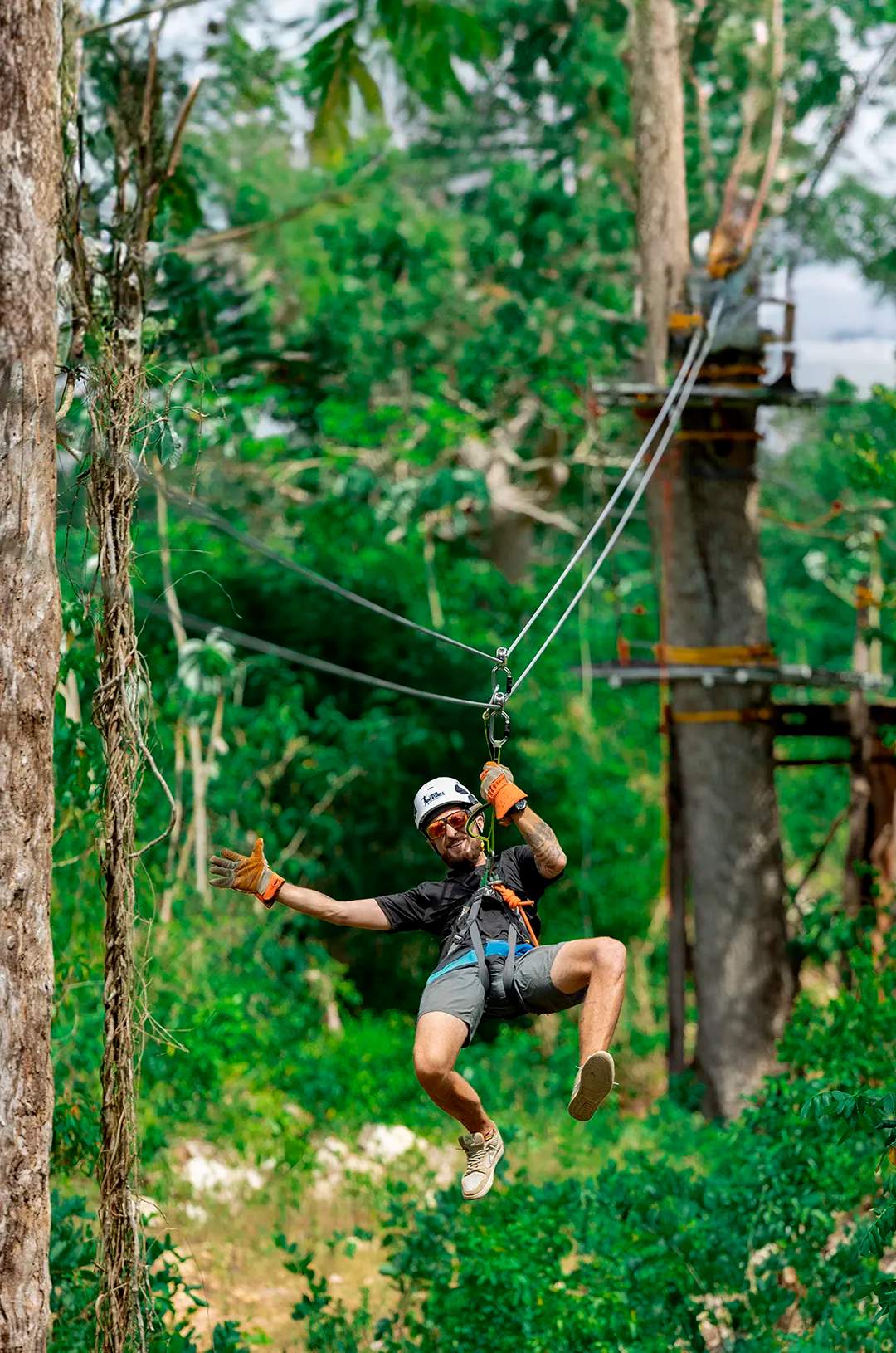 Un hombre haciendo tirolesa por un bosque, con casco y guantes, dando un pulgar arriba con una sonrisa.