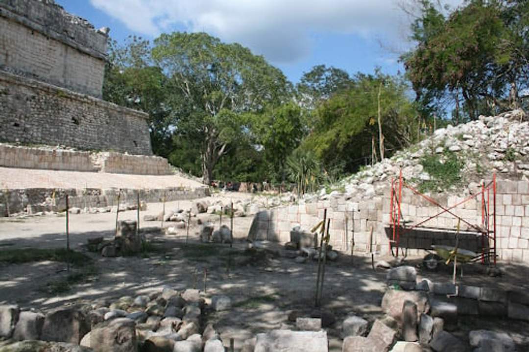 Sitio de excavación arqueológica con ruinas de piedra y rocas dispersas, parcialmente rodeado de árboles y vegetación. Andamios y equipos indican trabajos de restauración o estudio en curso.