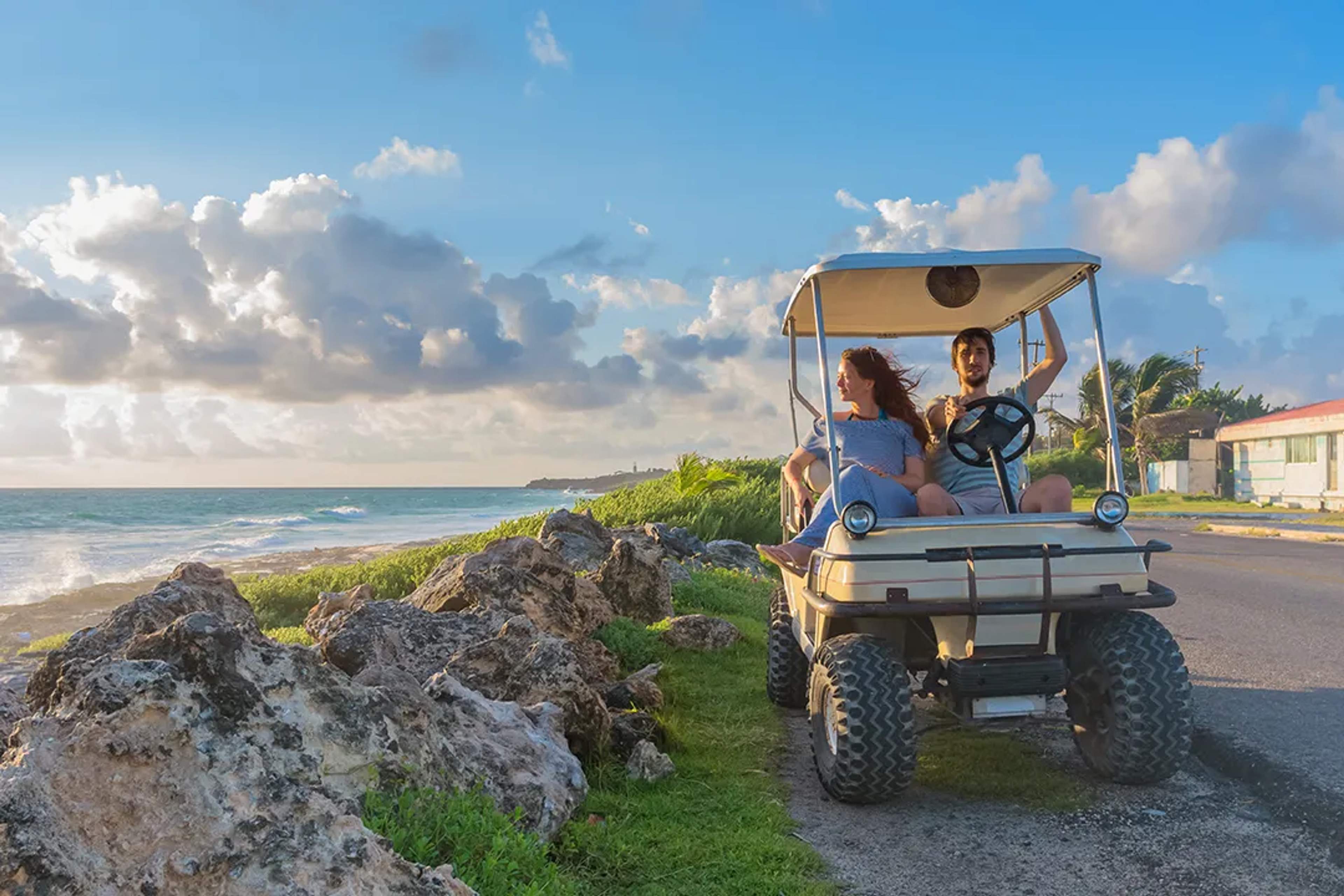 Couple enjoying a scenic coastal golf cart tour by the ocean under a sunny, cloudy sky at Playa Norte