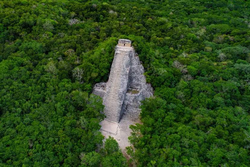Aerial view of a stone pyramid surrounded by dense green jungle, likely part of an ancient archaeological site.