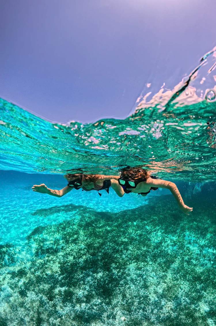Dos personas haciendo snorkel en aguas turquesas claras, con coral visible bajo el agua.