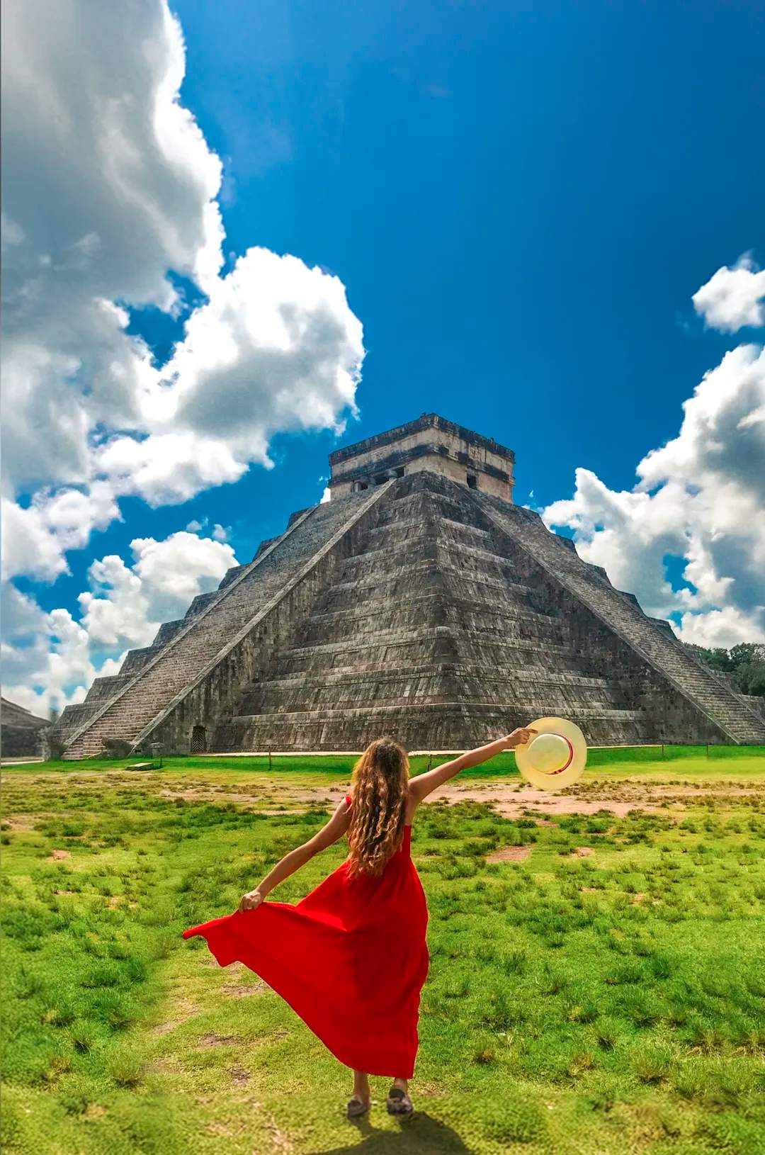 Woman in a red dress holding a hat, walking towards the Chichen Itza pyramid under a vibrant blue sky with scattered clouds.