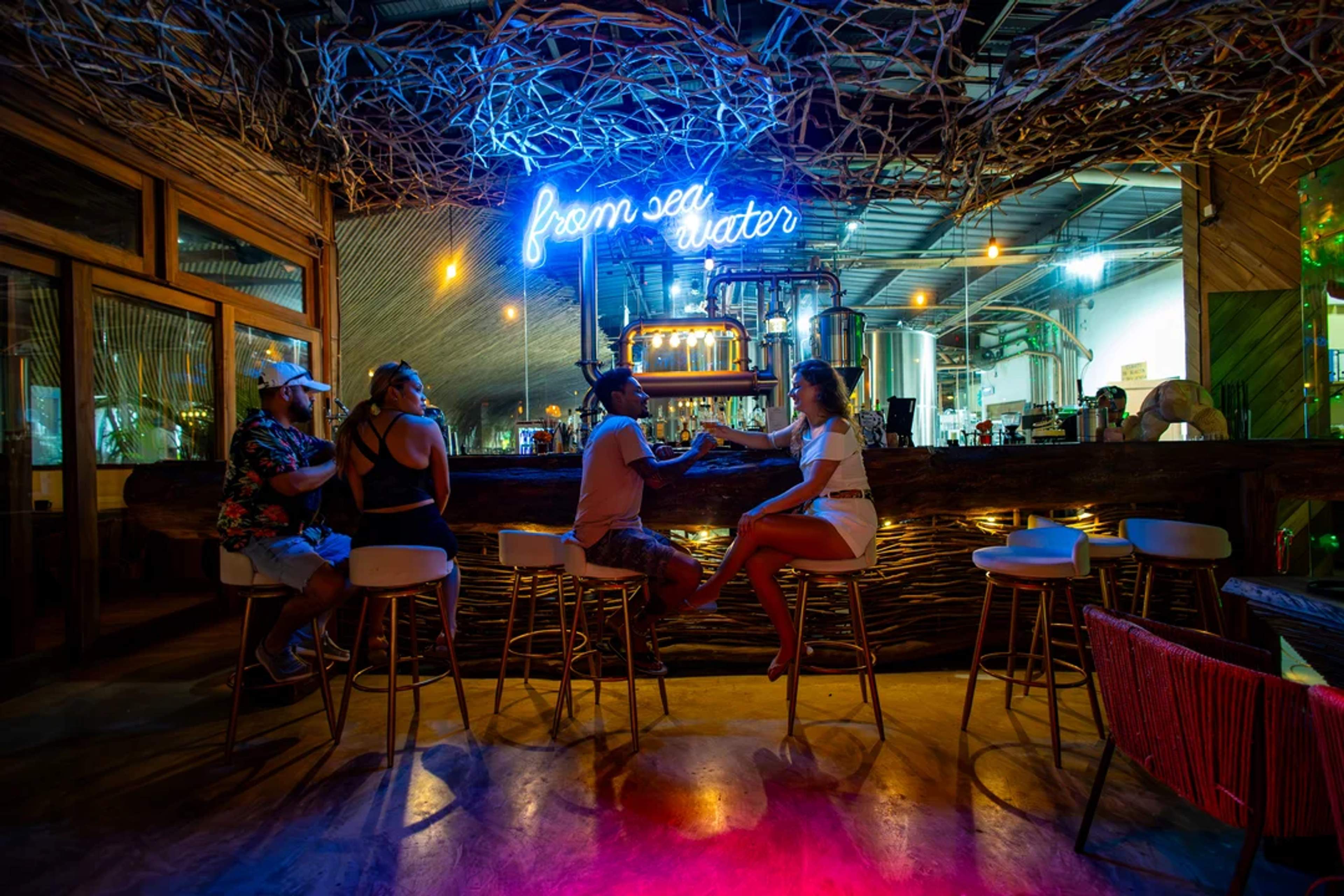 Parejas disfrutan tragos en un bar con luces neón y ambiente tropical estilo cervecería.