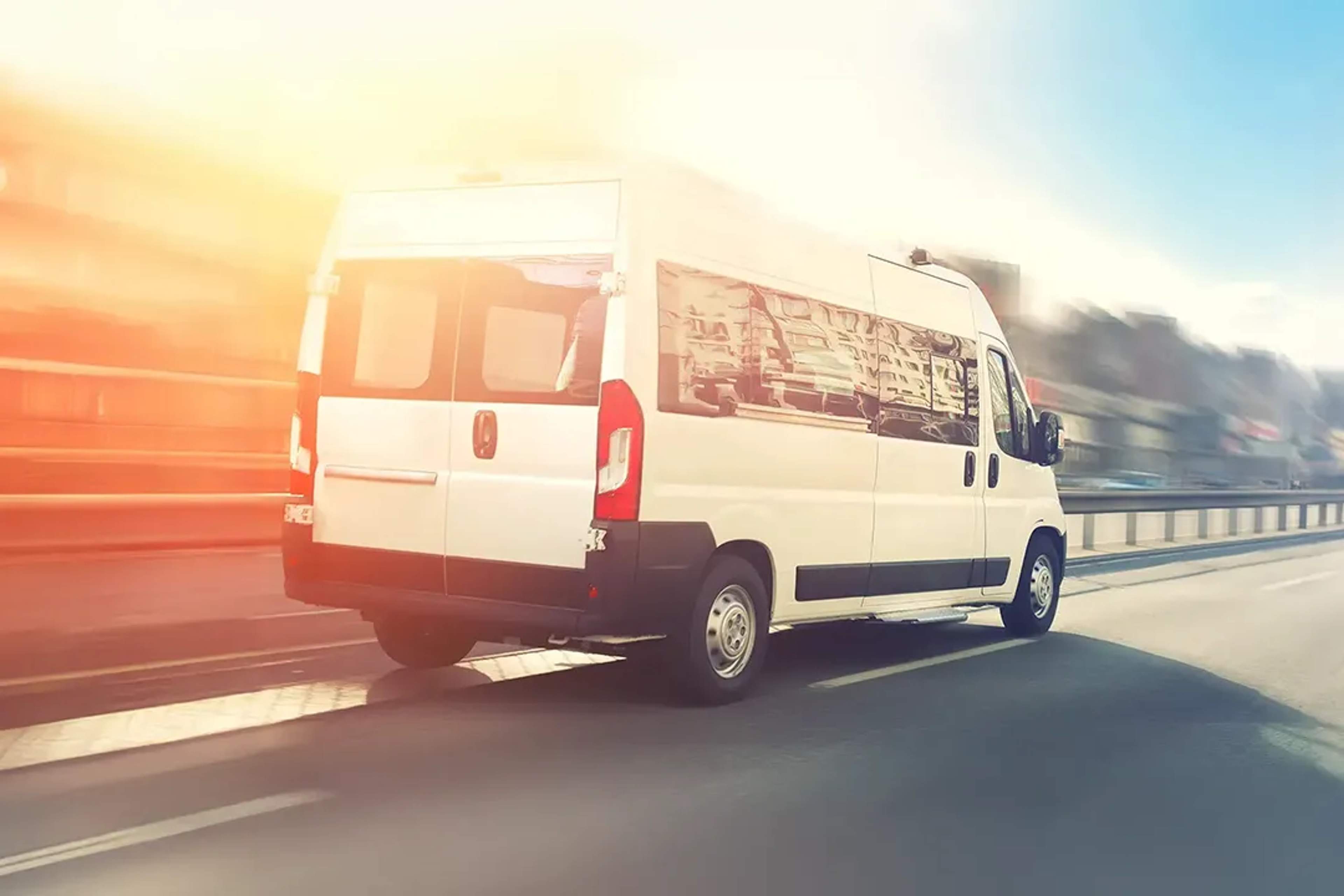 White passenger van driving on a highway at sunset, reflecting city buildings on windows.