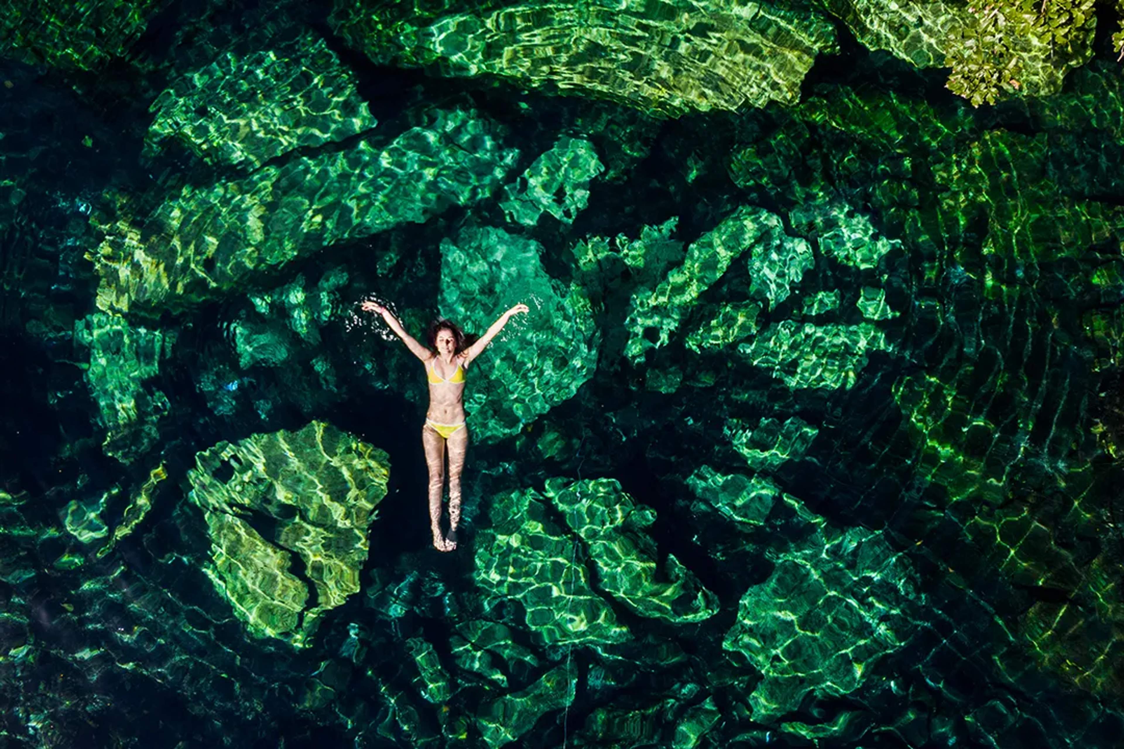 Woman floating in crystal-clear emerald cenote waters surrounded by rock formations in Riviera Maya nature tour