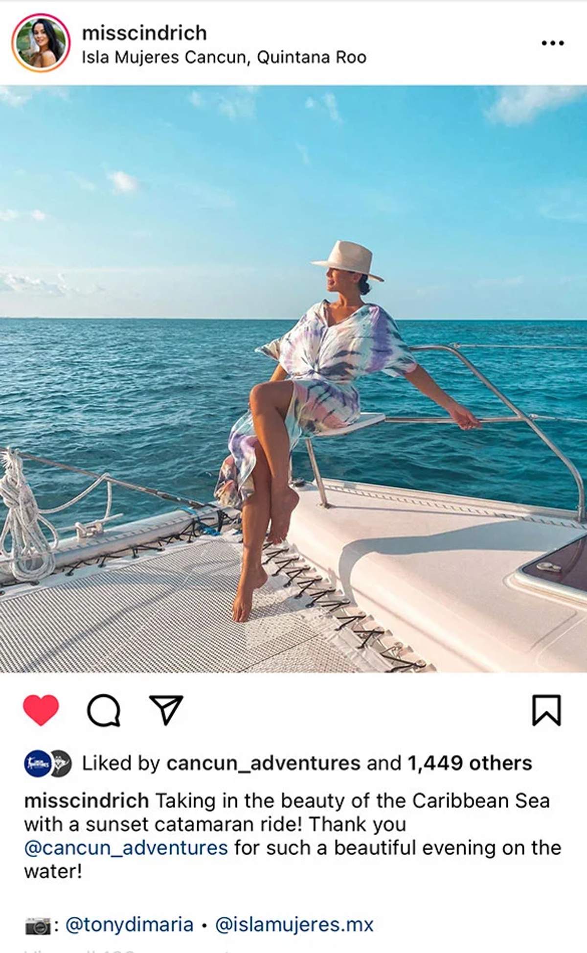 Spanish Alt Text: Una mujer con un vestido teñido y sombrero se sienta en el borde de un catamarán, disfrutando del Mar Caribe cerca de Isla Mujeres, Cancún al atardecer.