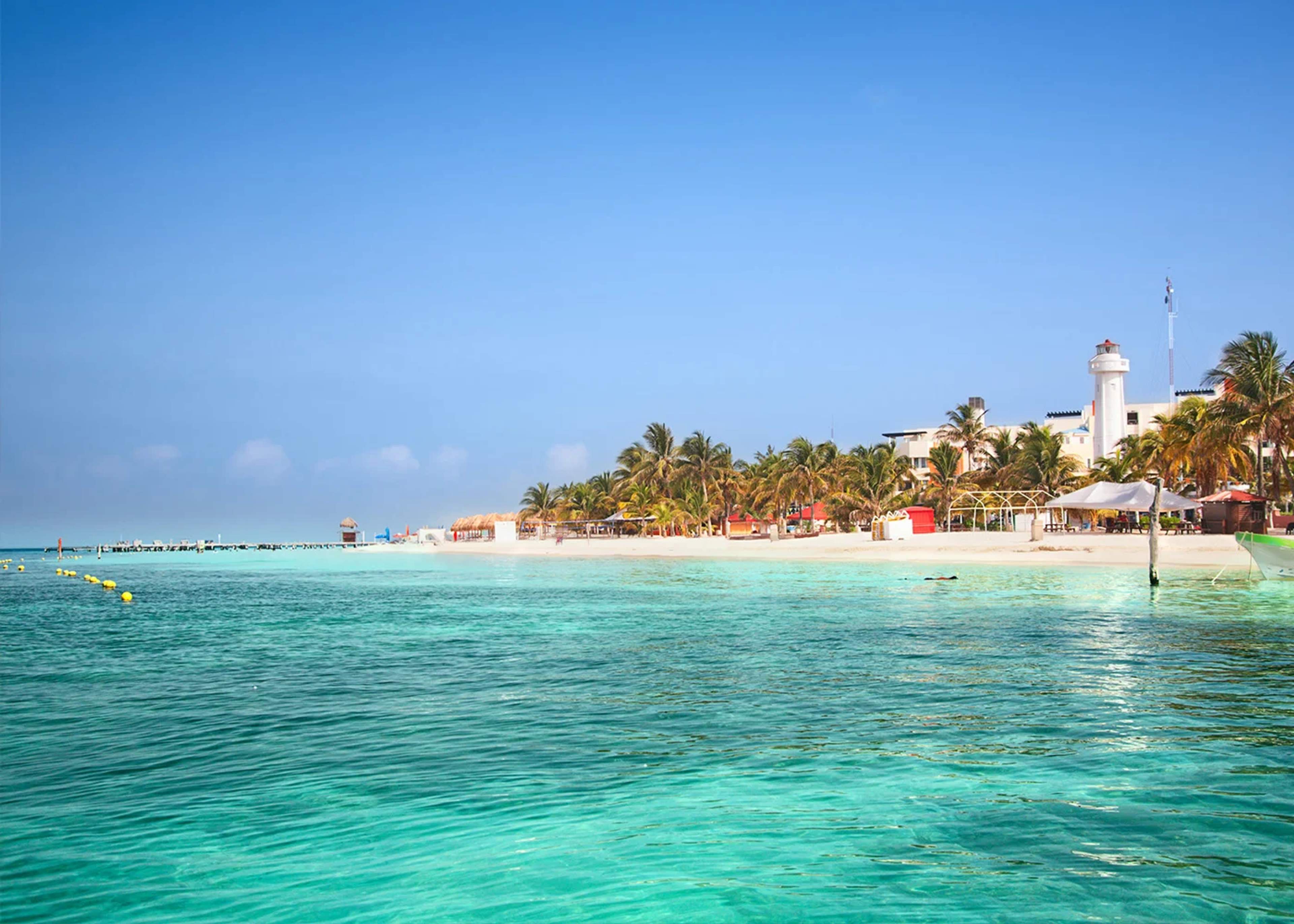 A beautiful beach scene with turquoise water and white sandy shores lined with palm trees. In the background, there are several buildings, including a lighthouse, under a clear blue sky.