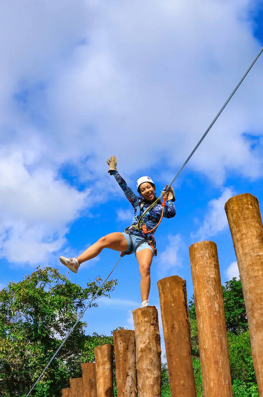 Una mujer equilibrándose en postes de madera con arnés de seguridad, sonriendo y levantando el brazo bajo un cielo azul.