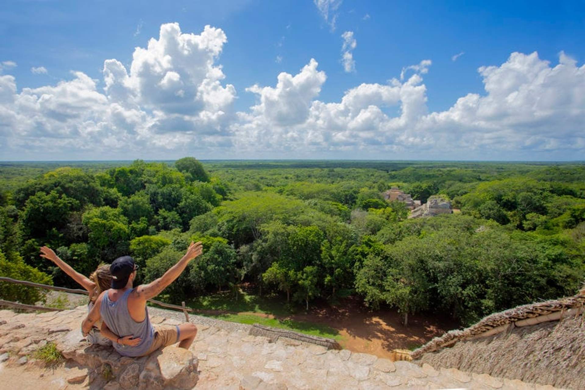 A couple sitting on the edge of a pyramid at Ek Balam, overlooking the lush green jungle under a bright blue sky with scattered clouds.