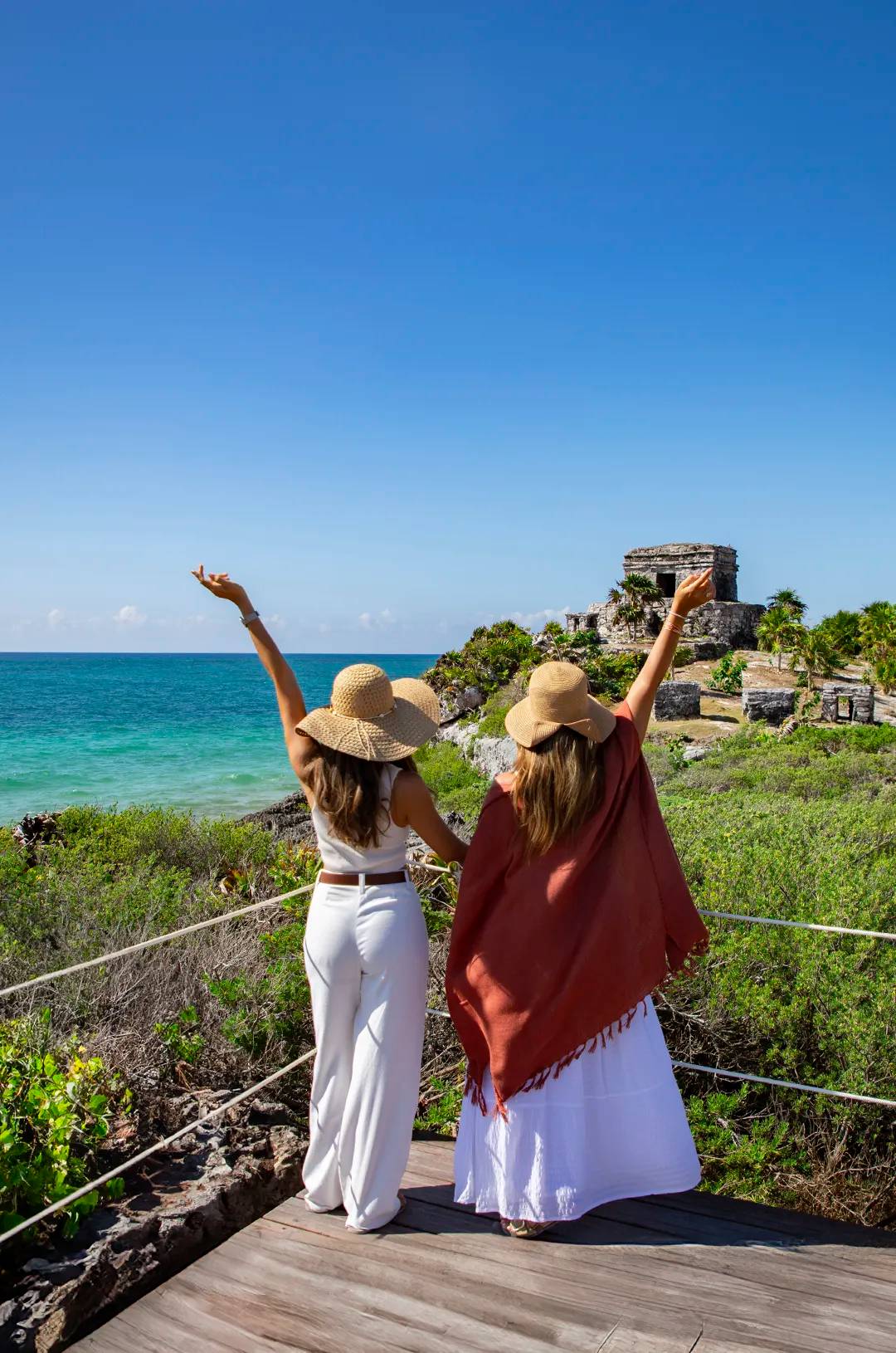 Two women in hats, backs turned, pose by the Tulum ruins with arms raised, overlooking the turquoise sea and clear sky.