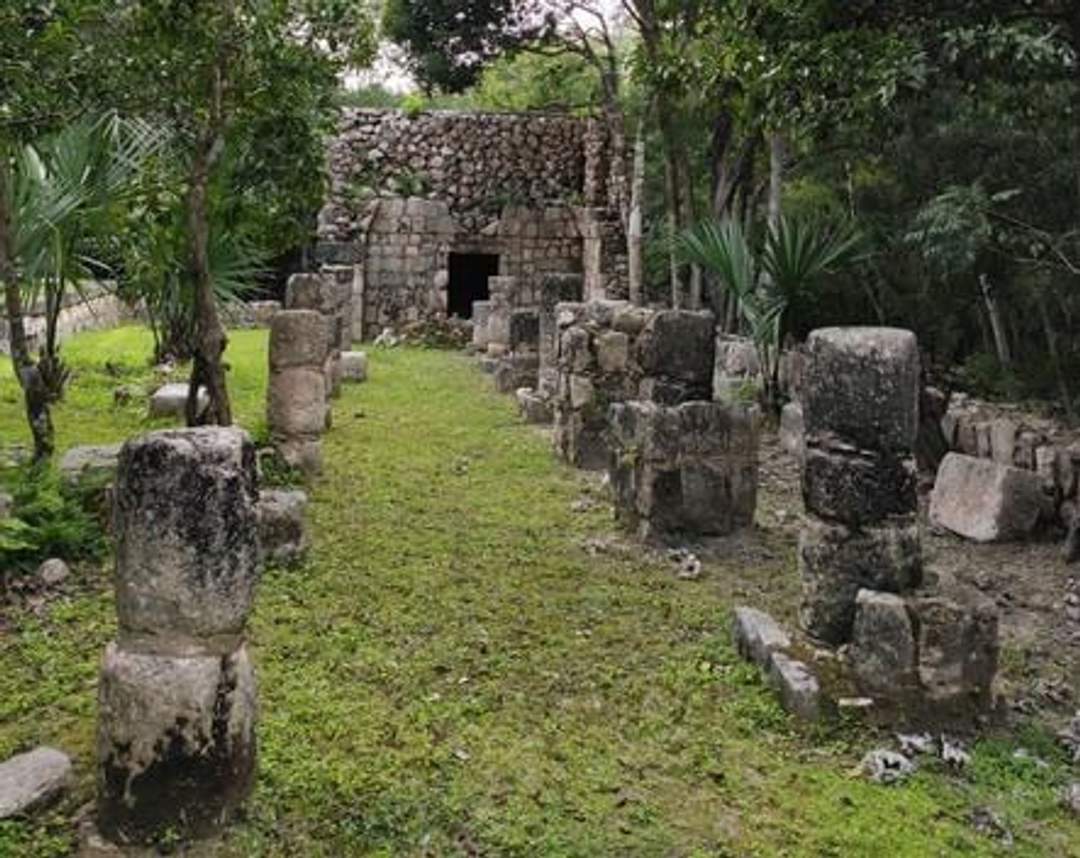 A photograph of the Gallery of the Monkeys at Chichen Itza, featuring an ancient stone structure with a row of columns leading to a small building with a doorway. The site is surrounded by lush v