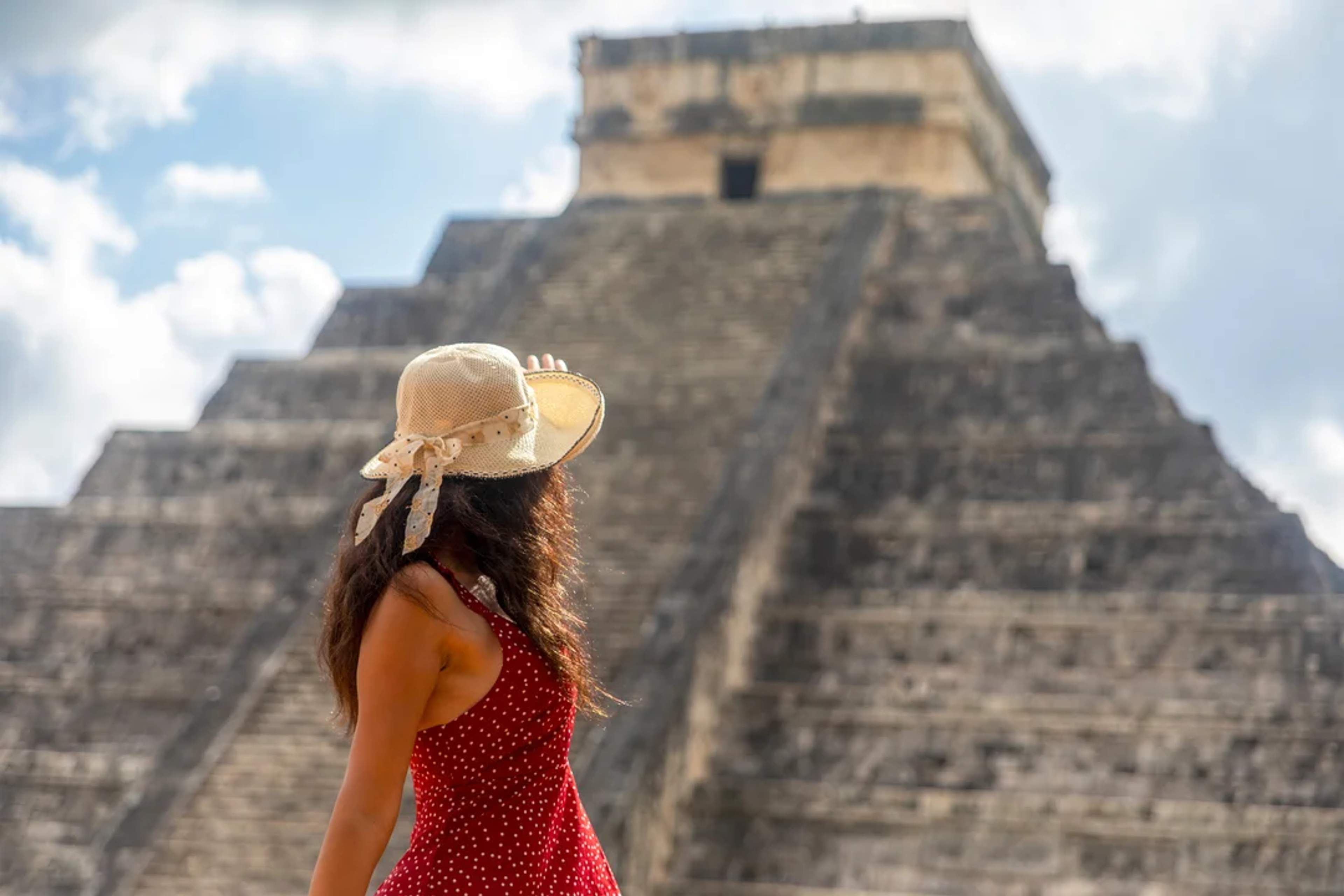Una mujer con sombrero y vestido rojo observa la pirámide de Chichén Itzá bajo un cielo parcialmente nublado.