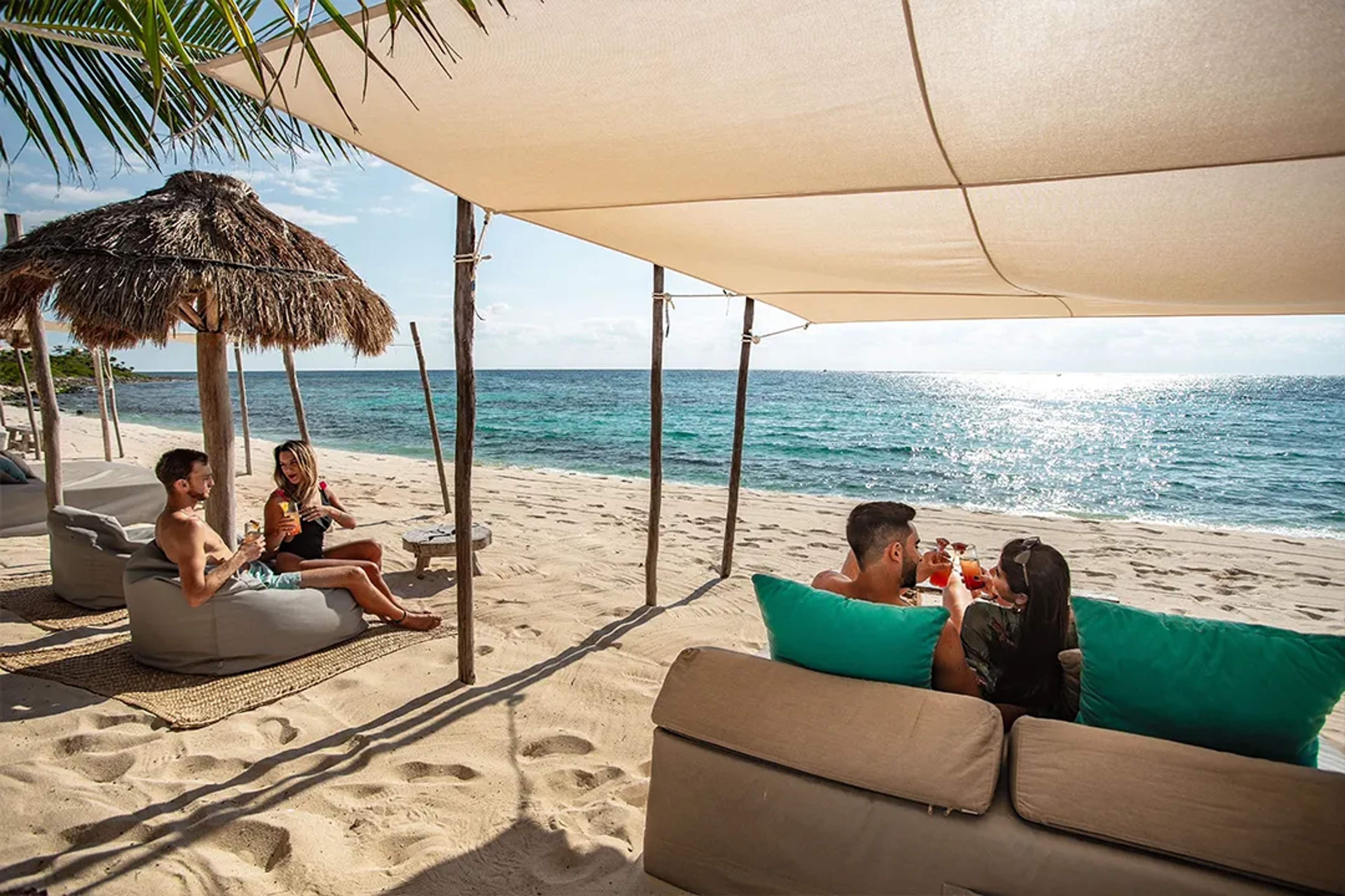 Couples enjoying cocktails on beachfront lounge beds under shade with Caribbean Sea views during luxury beach club day