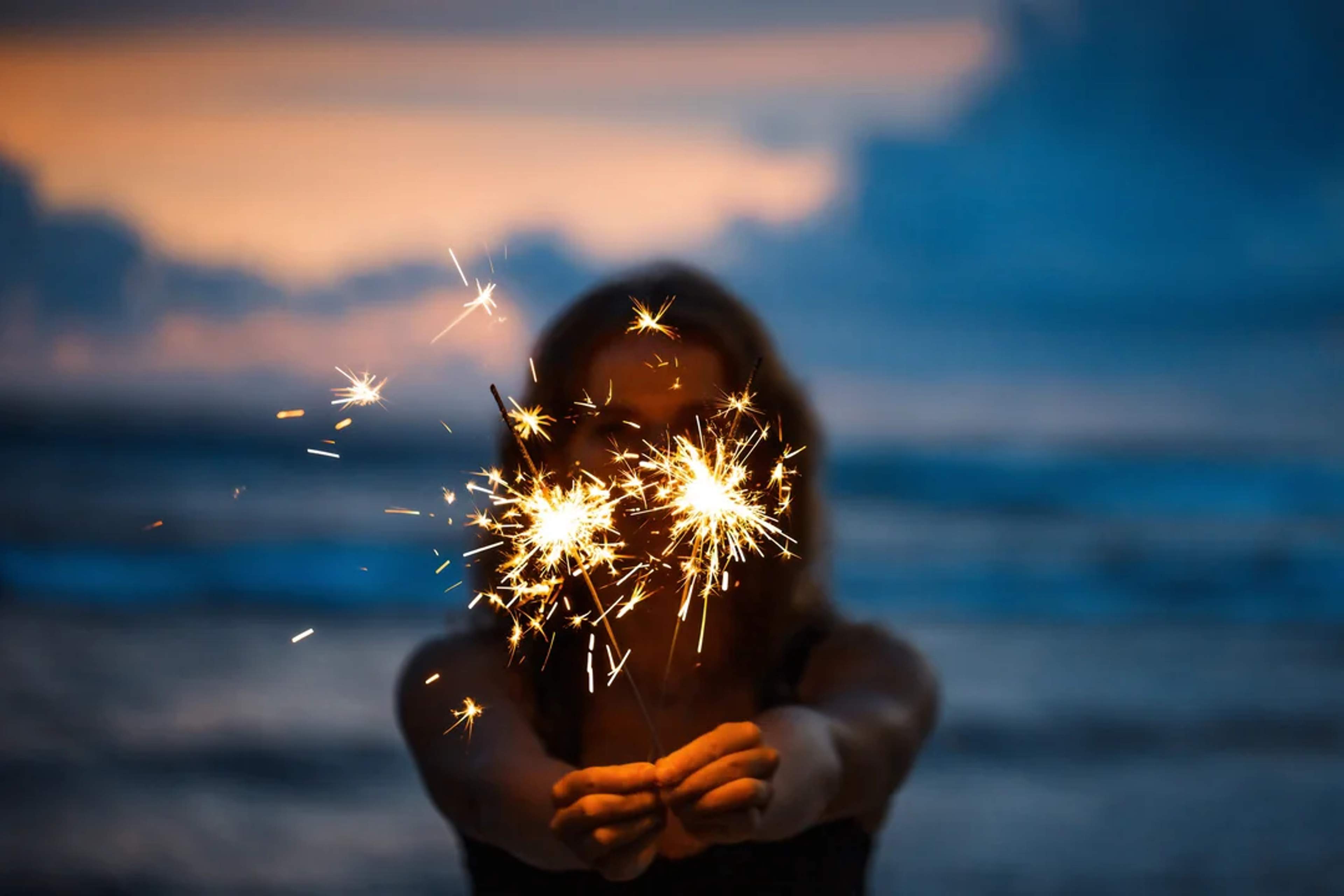 Woman holds sparklers at the beach during sunset, creating a festive and magical moment.