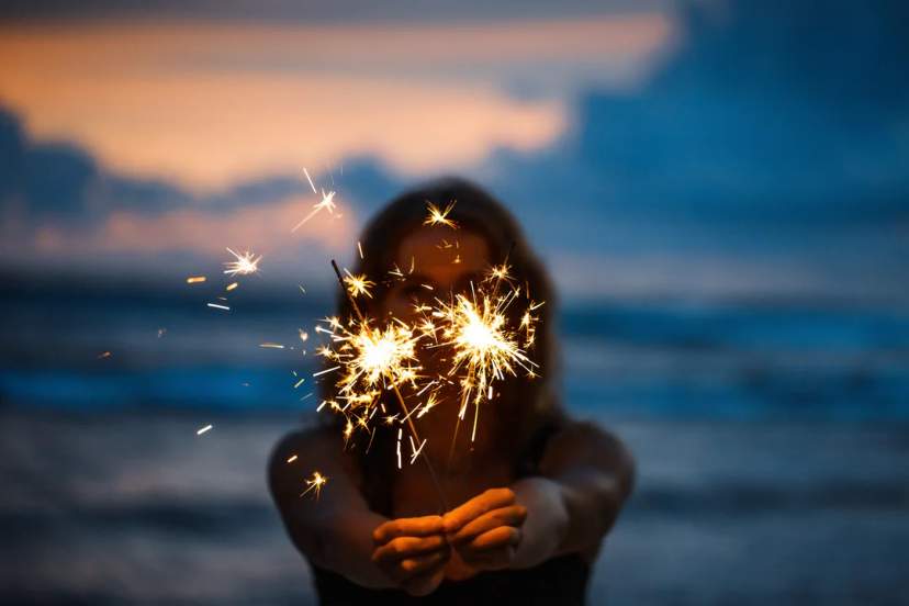 Mujer sostiene bengalas en la playa al atardecer, creando un momento festivo y mágico.