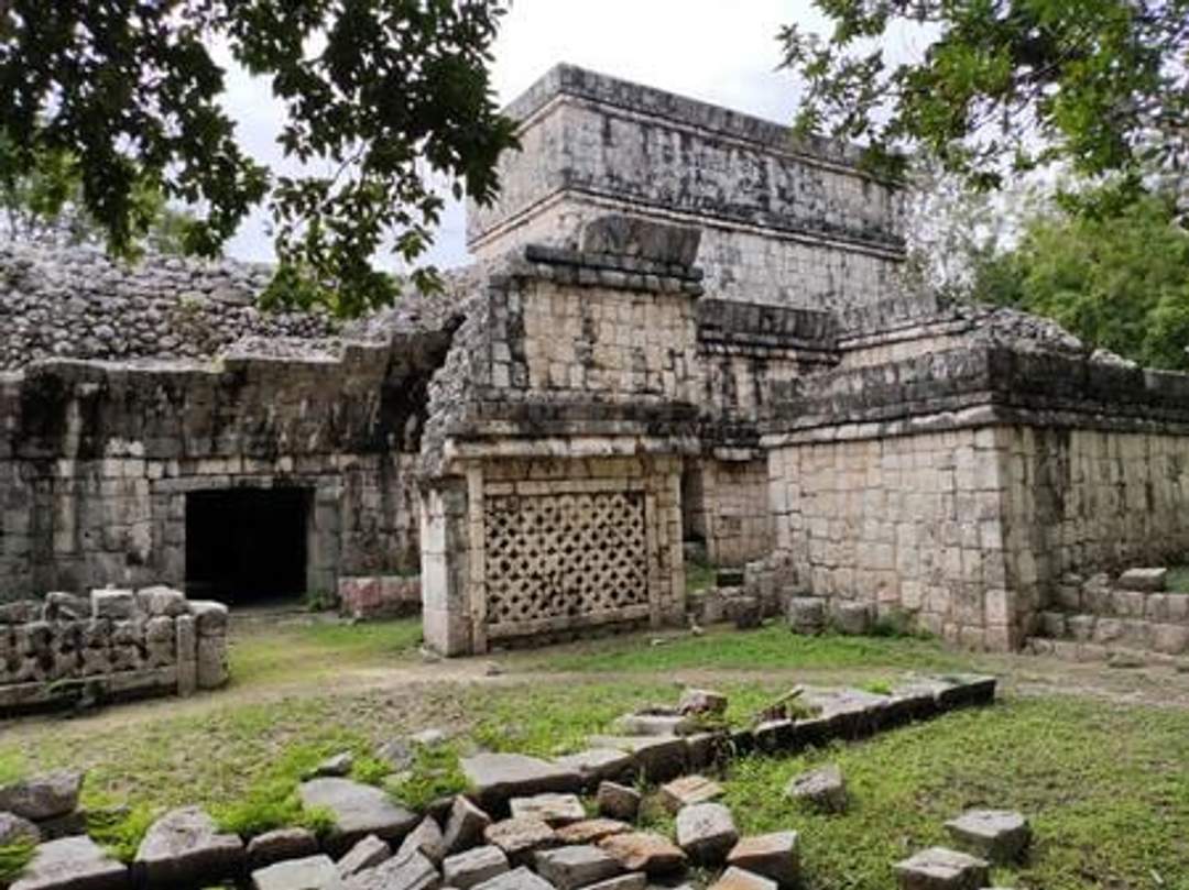 A photograph of the House of the Phallus at Chichen Itza, featuring a complex of ancient stone structures with various decorative elements, including a lattice-like stonework. The ruins are surrounded by greenery. Photo credit: Giovanni Agostino Frassetto.