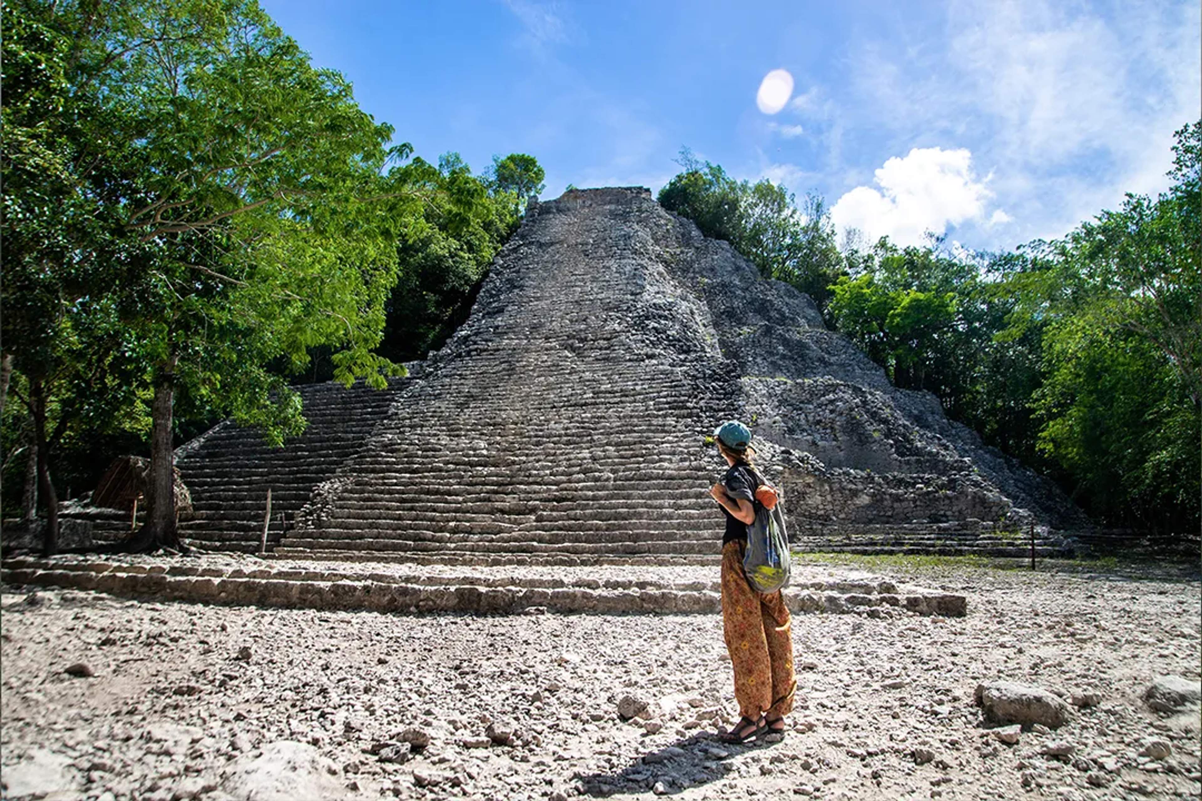 Tourist admiring the ancient pyramid of Cobá, surrounded by jungle under a bright sunny sky.