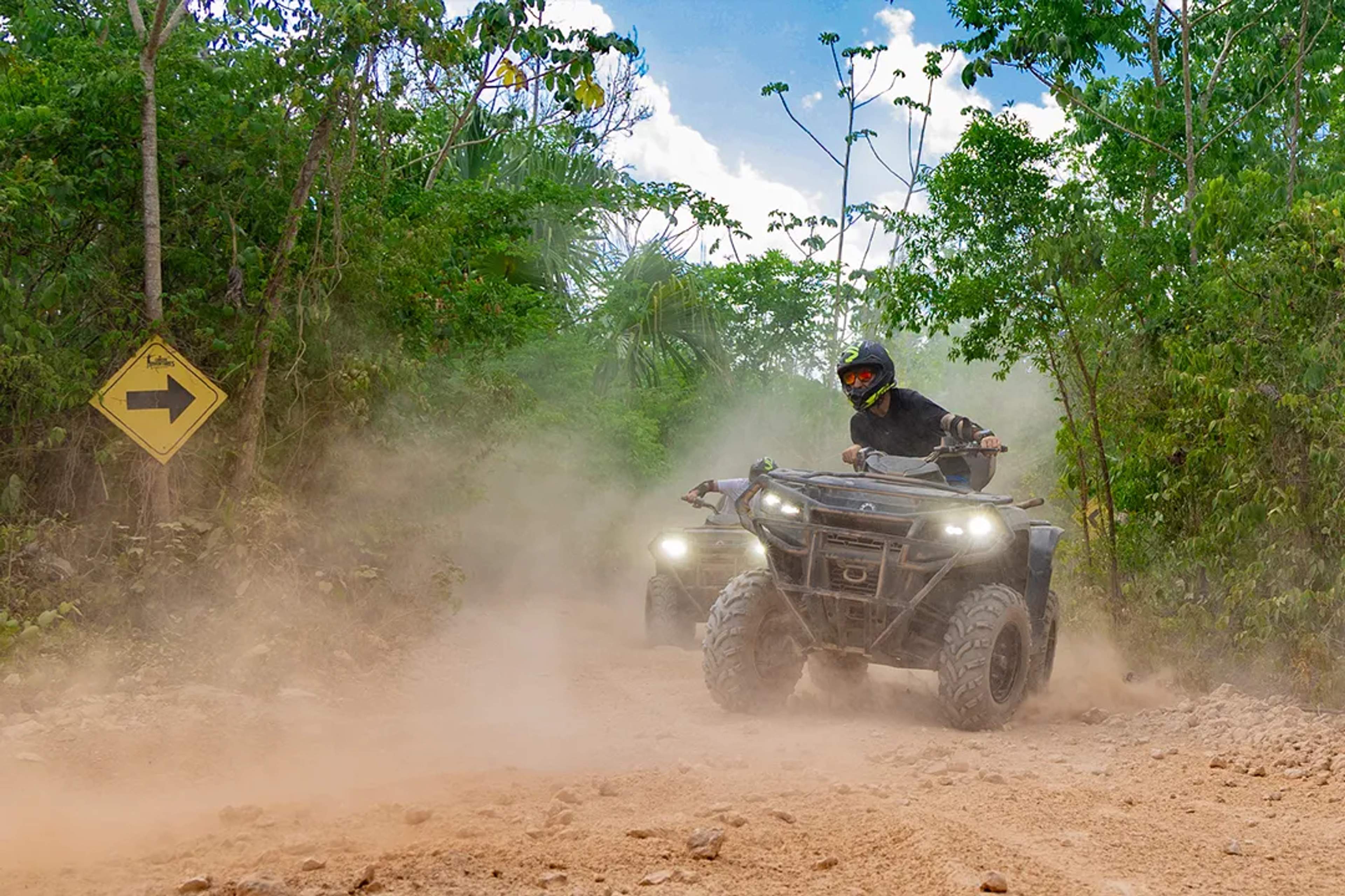 Dos conductores en cuatrimotos recorren un camino polvoso en la selva, rodeados de vegetación.