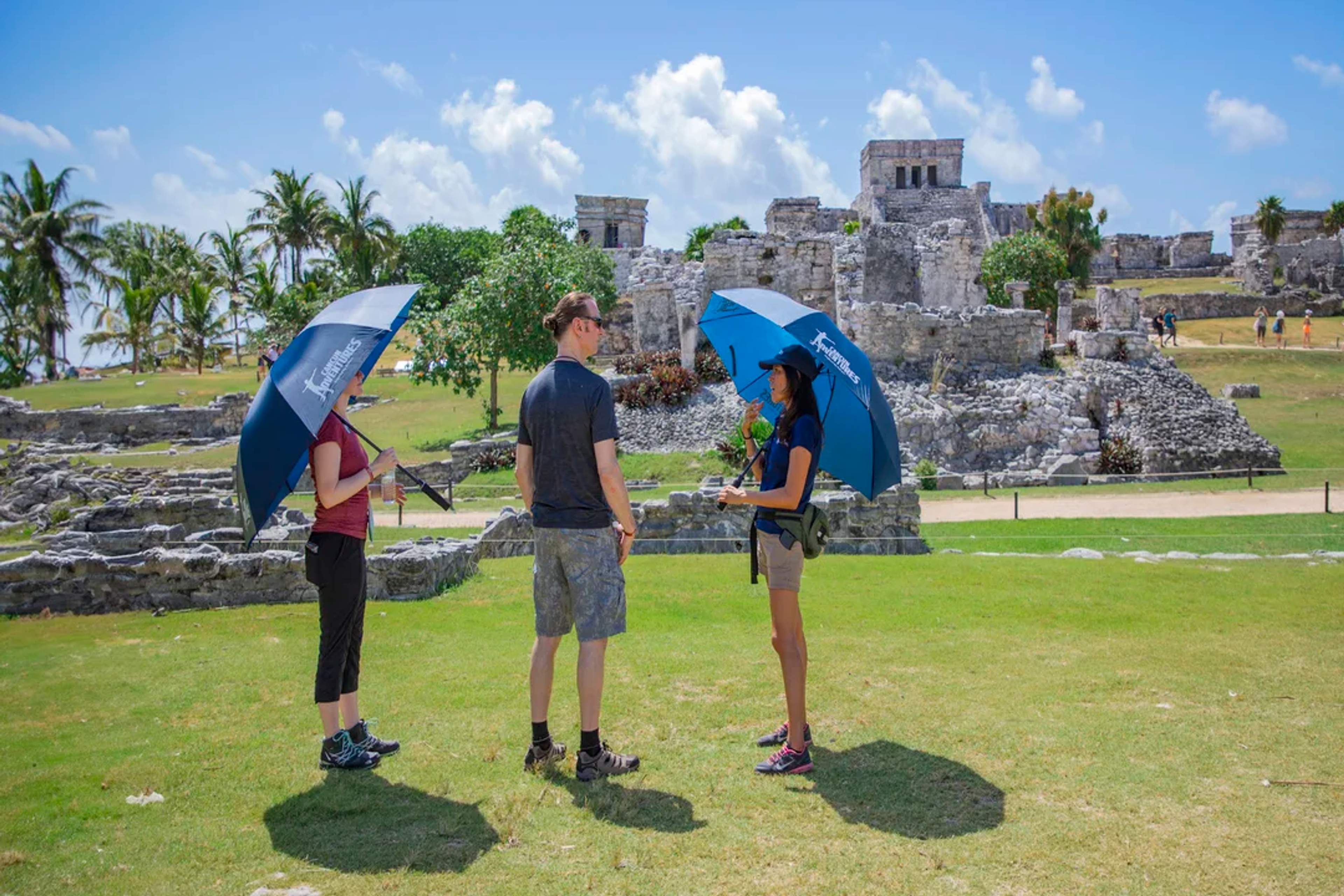 Guided tour in Tulum ruins under sunny skies, guests stay cool with branded umbrellas.