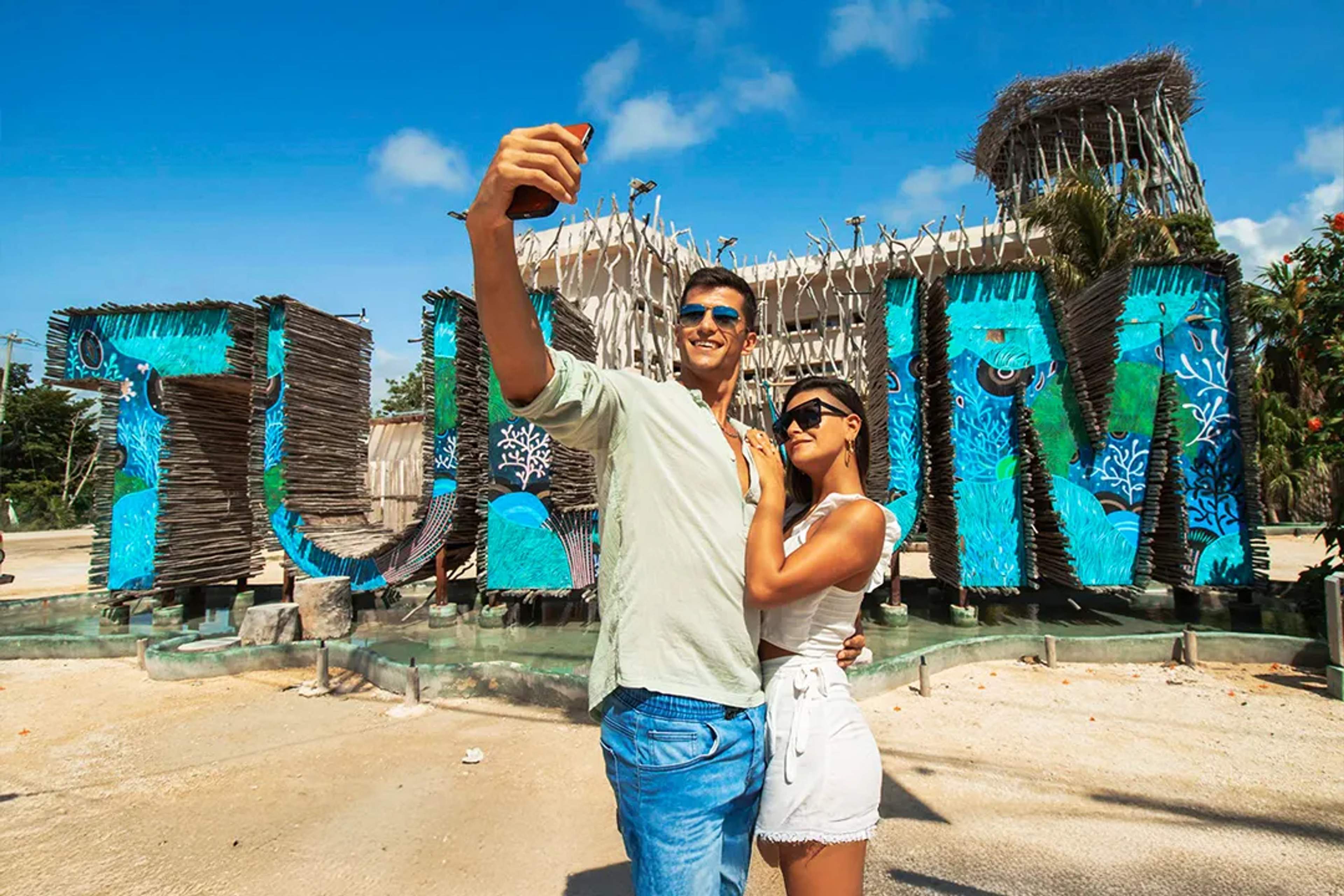 Couple taking a selfie at Tulum sign in Riviera Maya with colorful artwork and tropical setting on a sunny day