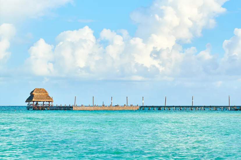 Pier with palapa over turquoise Caribbean water in Cancún under bright sky and soft white clouds.