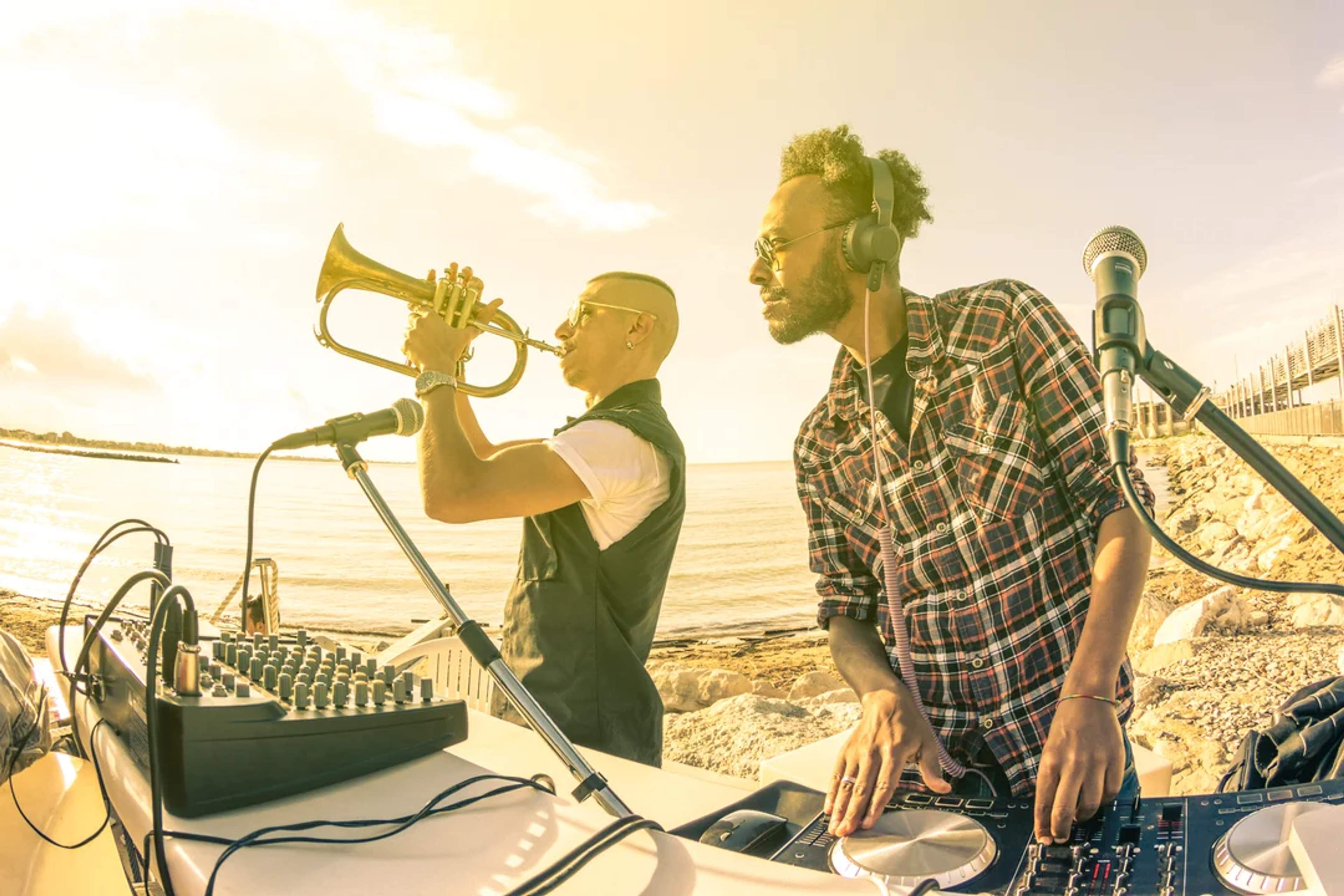 DJ and trumpet player perform by the sea, blending music and sunset vibes outdoors.