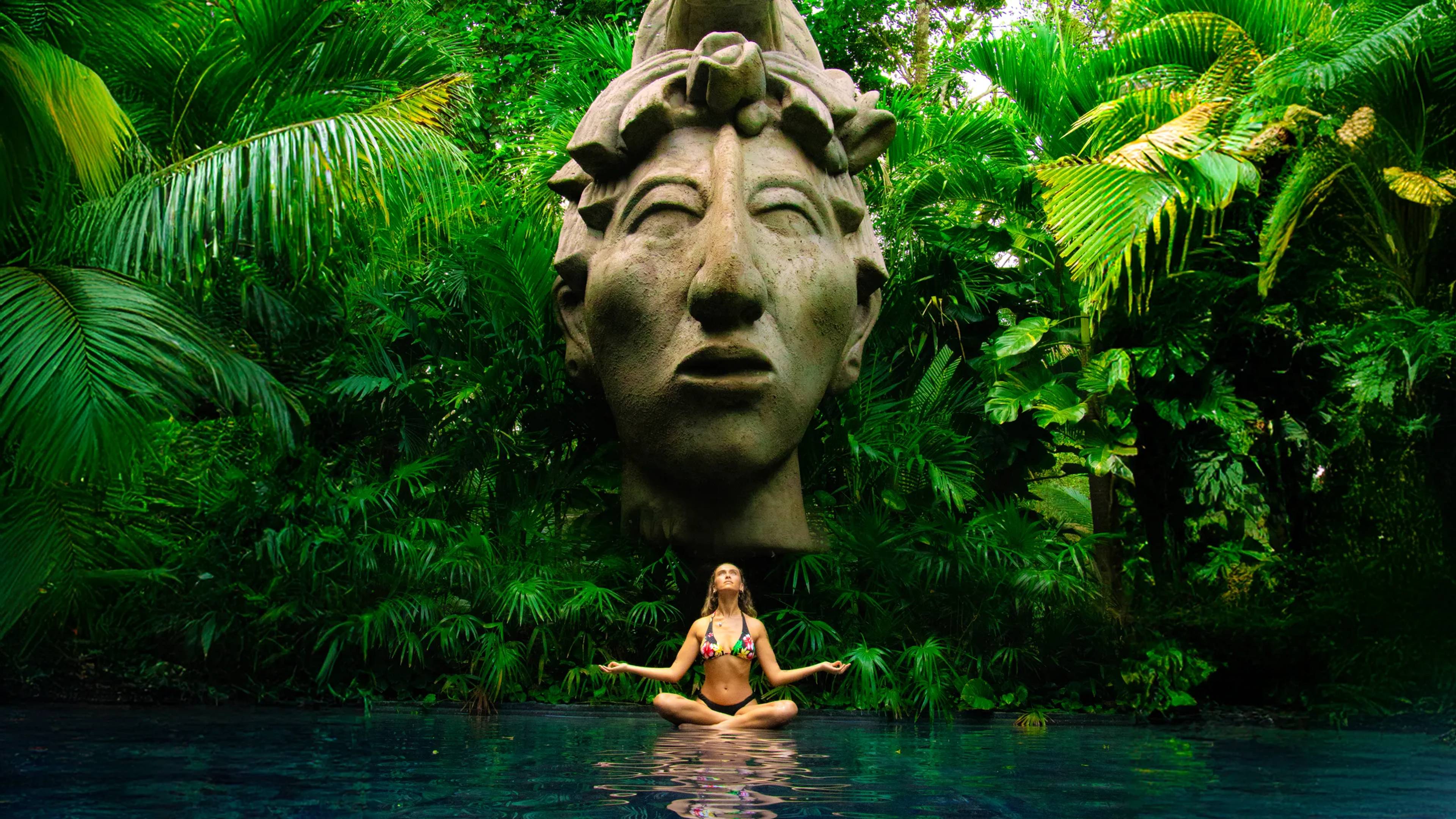 Mujer meditando en una piscina con una escultura maya gigante en la exuberante vegetación de Cancún.